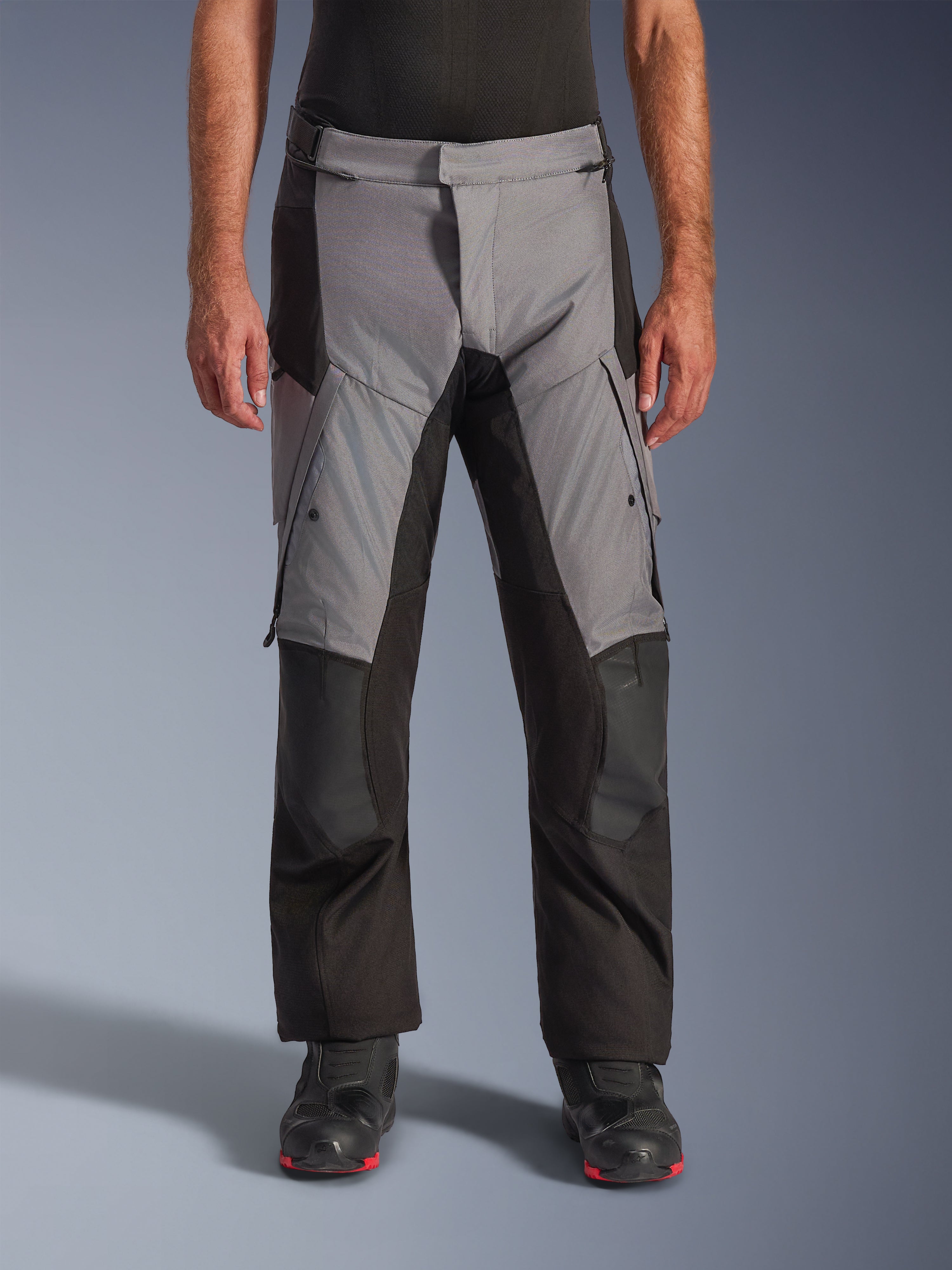 Alpinestars Andes V4 Drystar® byxor, adventure touring-byxor för motorcykel, grå och svarta, framifrån buren av en modell med svarta mc-stövlar, med förstärkta knäpartier och cargofickor på låren, vattentät och andningsbar textilkonstruktion för körning i alla väder