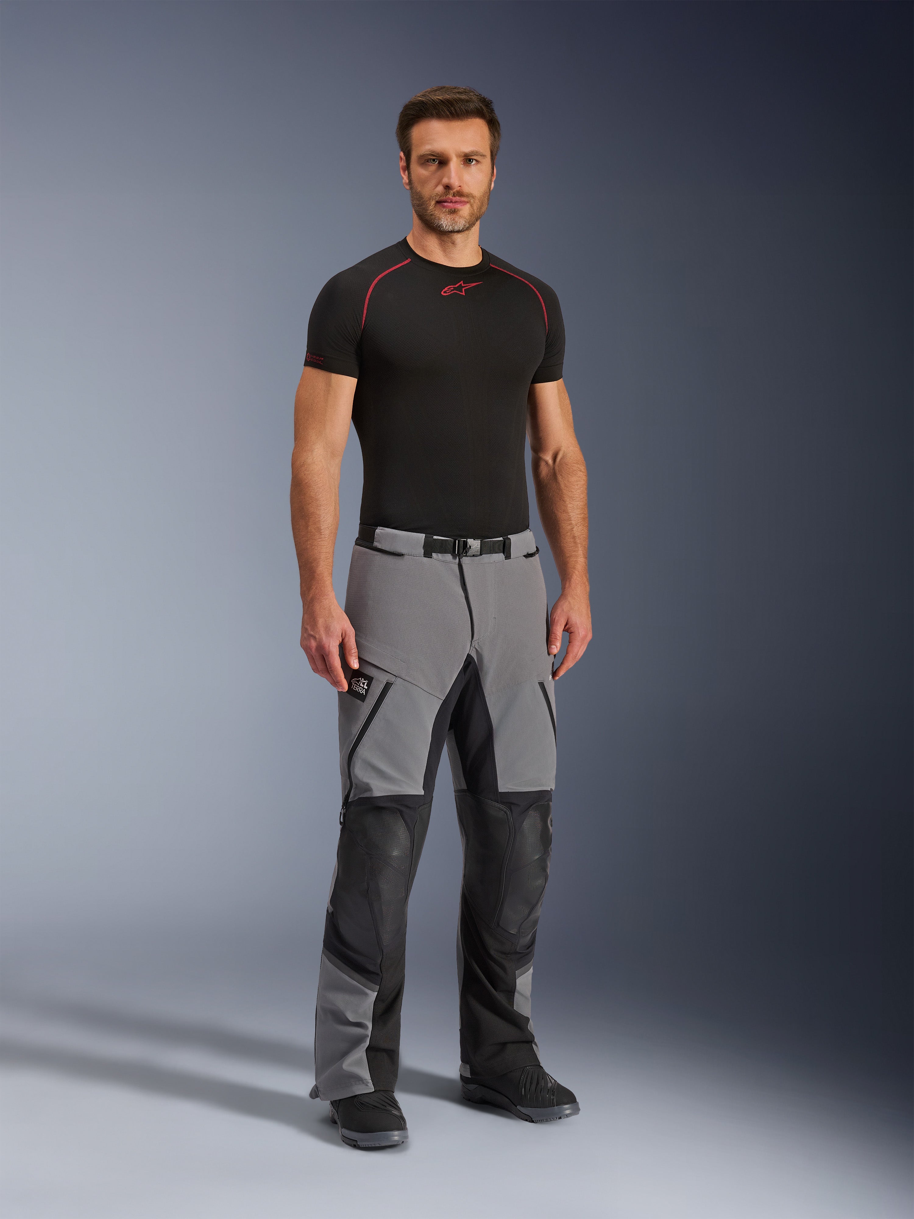 Alpinestars Andes Pro Drystar® XF byxor, äventyrsbyxor för motorcykel, mörkgrå och svarta, bärs av en modell med ett svart tekniskt underställ, med flerpanelskonstruktion, vattentäta dragkedjor och förstärkta zoner för äventyrskörning.