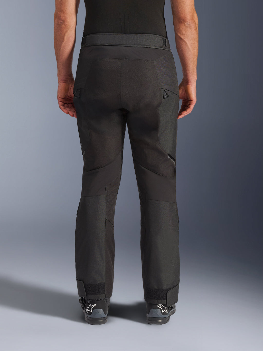 Alpinestars Cusco Drystar® byxor, MC-byxor, svarta, sedda bakifrån burna av en förare, med texturerade textilpaneler, ventilationsöppningar med dragkedja på låren och justerbara benslut, kombinerade med svarta Alpinestars-stövlar.