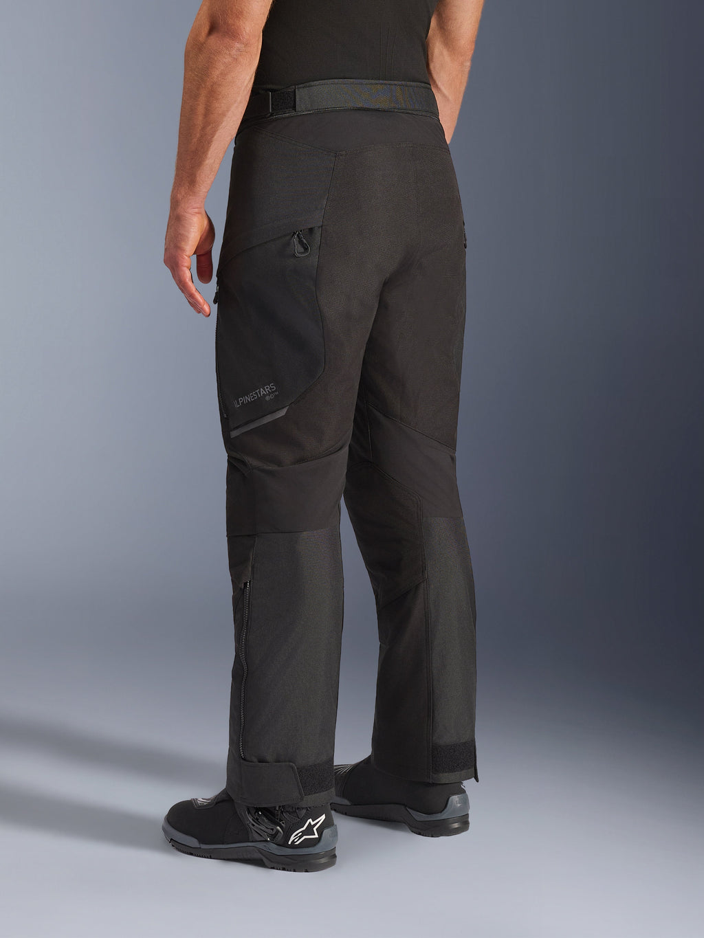 Alpinestars Cusco Drystar® byxor, motorcykelbyxor, svarta, sedda snett bakifrån med lårficka med dragkedja och artikulerade paneler, bärs med svarta motorcykelstövlar, hållbar adventure touring-design med diskret logotyp