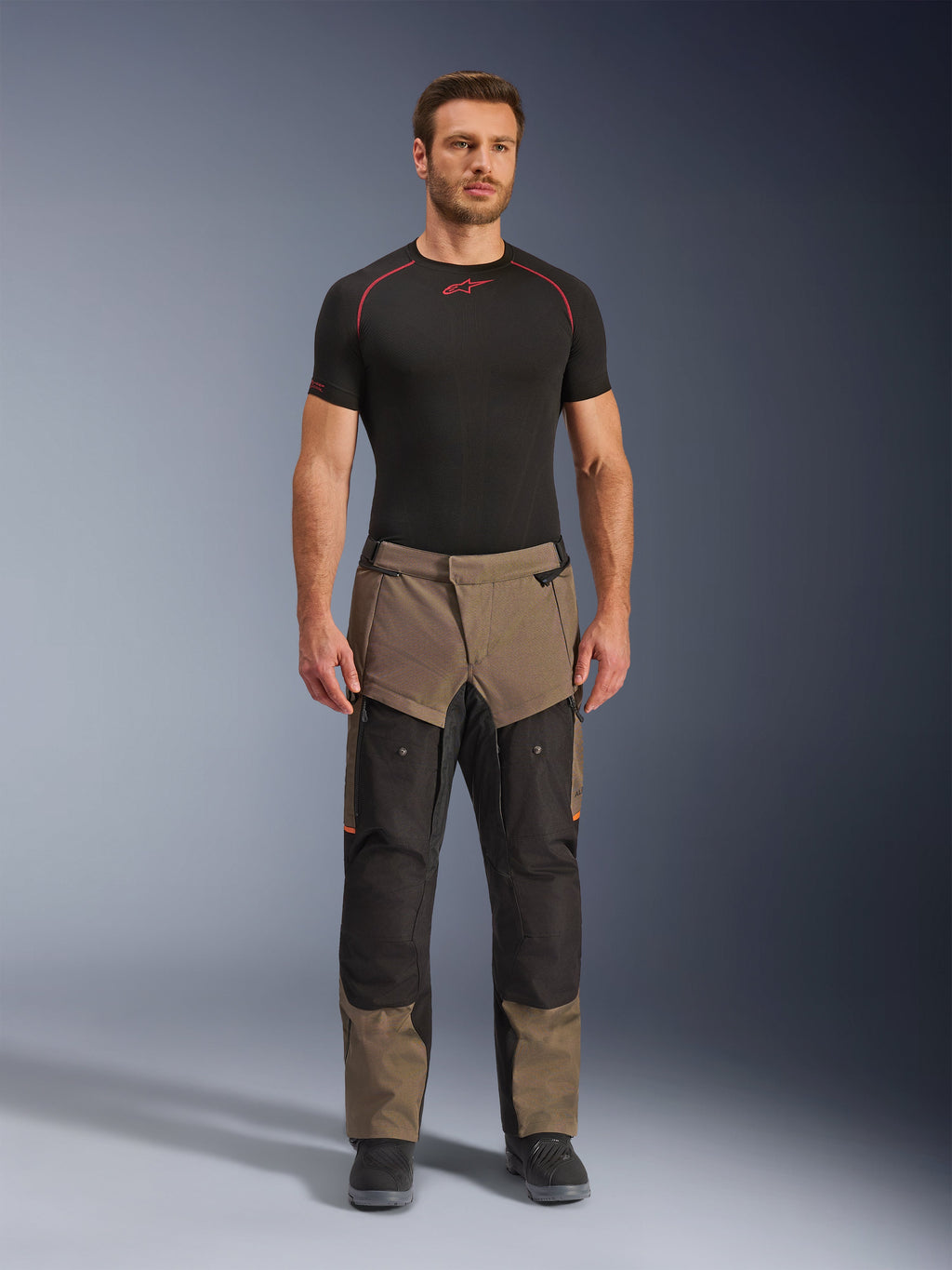 Alpinestars Cusco Drystar®-byxor, Walnut Black, adventure touring-motorcykelbyxor, bärs av en manlig modell med svart underställ och svarta stövlar, med valnötsbruna och svarta paneler, orangea detaljer och direkta ventilationsdragkedjor på låren
