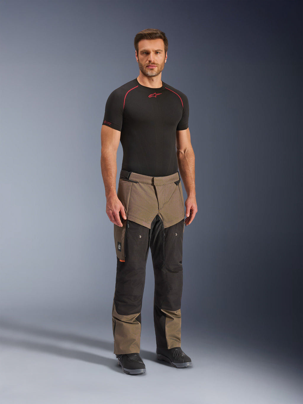 Alpinestars Cusco Drystar® Pants, adventure-motorcykelbyxor, valnöt och svart, bärs av en modell med en svart underställströja och stövlar, med stora benfickor på låren, ventilationsdragkedjor och en robust tvåfärgad design.