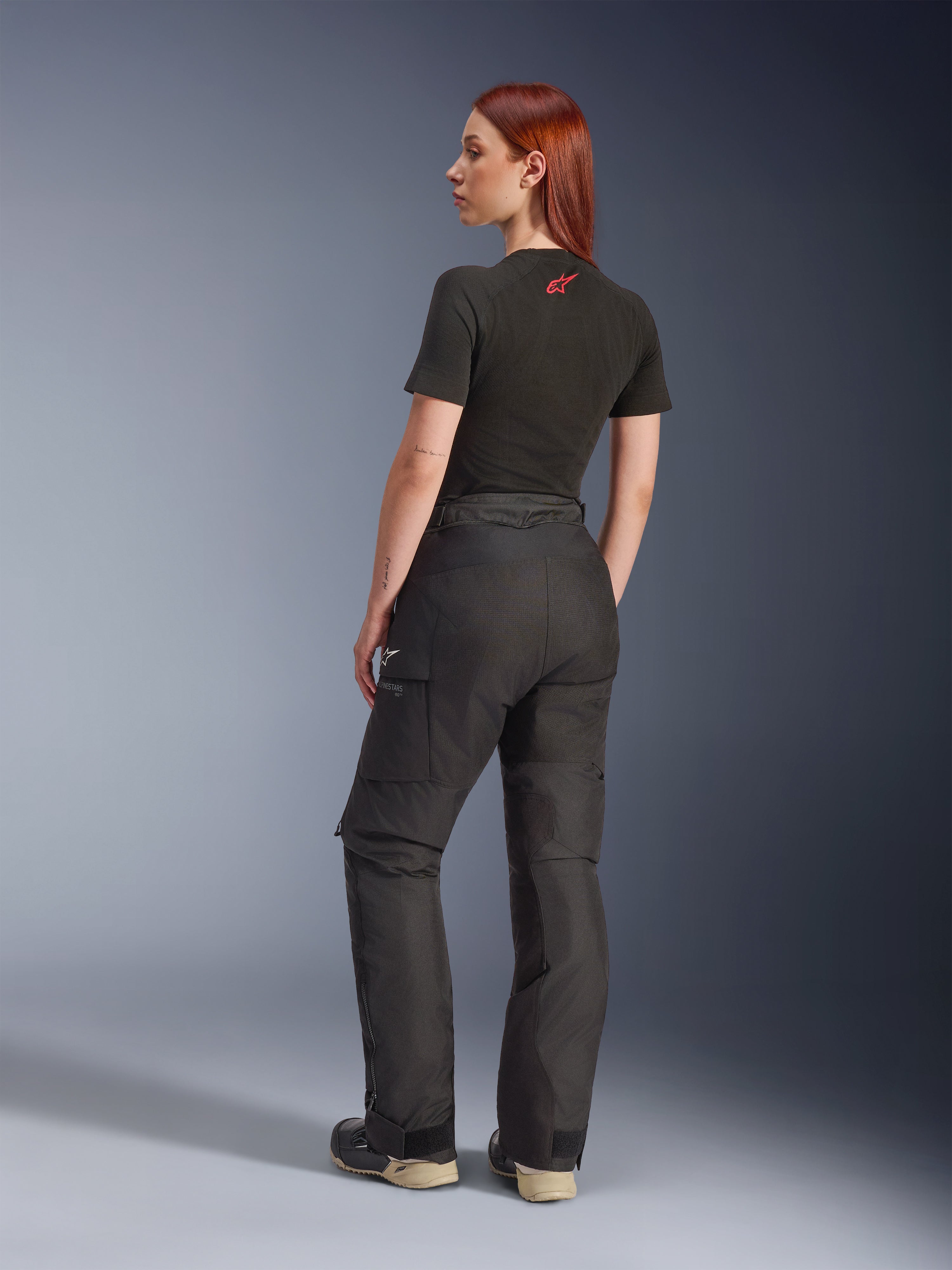 Alpinestars Stella Andes V4 Drystar®-byxor, svarta, äventyrsmotorcykelbyxor som visas från en vinkel snett bakifrån på en modell, kombinerade med en svart t-shirt och stövlar, med cargofickor på sidorna med vit logotyp och hållbar textilkonstruktion.