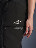 Alpinestars Stella Andes V4 Drystar® byxor, motorcykelbyxor för dam, svarta, närbild på cargoficka på låret med vit stjärnlogotyp och märkesnamn, hållbart textilmaterial för äventyrskörning