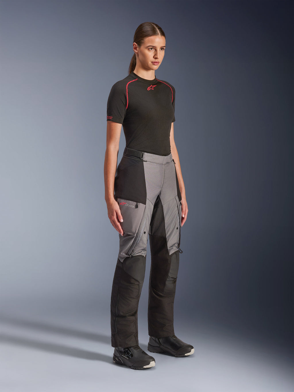 Alpinestars Stella Andes V4 Drystar® byxor, äventyrsbyxor för damer, svart och mörkgrå, bärs av en modell med ett svart Alpinestars-underställ och svarta stövlar, med förstärkta paneler, röd Drystar-logotyp och cargofickor.