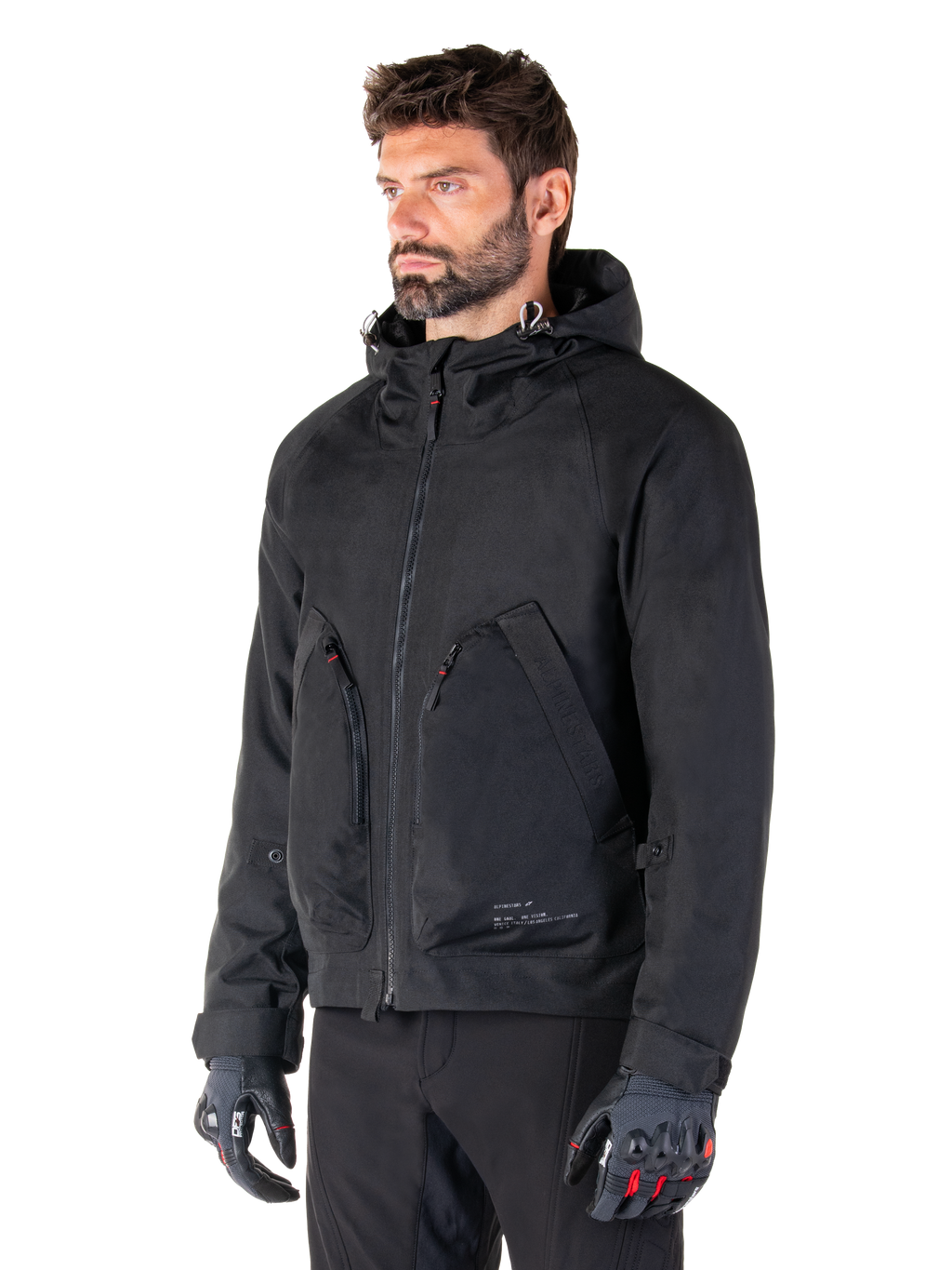 Man wearing Alpinestars Morush Waterproof Parka, Black urban motorcycle jacket, streetwear-inspired design with hood and large zippered front pockets, paired with grey and black motorcycle gloves, contemporary minimalist style