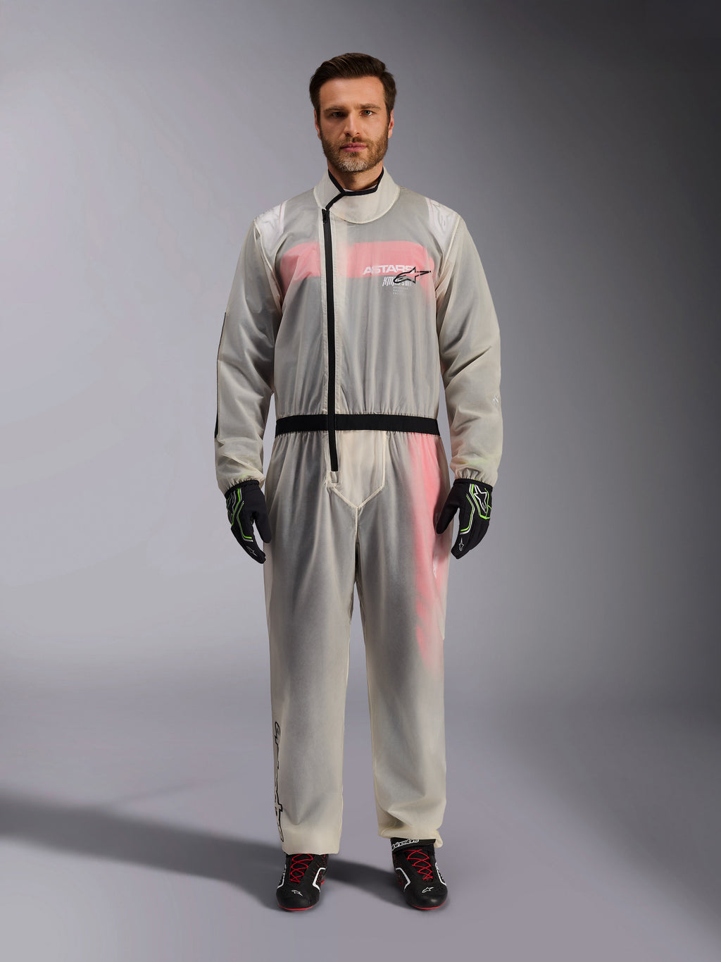 Alpinestars Kart Rain Suit, Clear, genomskinligt vattentätt regnställ som bärs av en modell, med en svart asymmetrisk dragkedja och logotyp på bröstet, röd och vit racingdräkt synlig under, matchat med svarta och neongröna handskar och svarta racingstövlar.