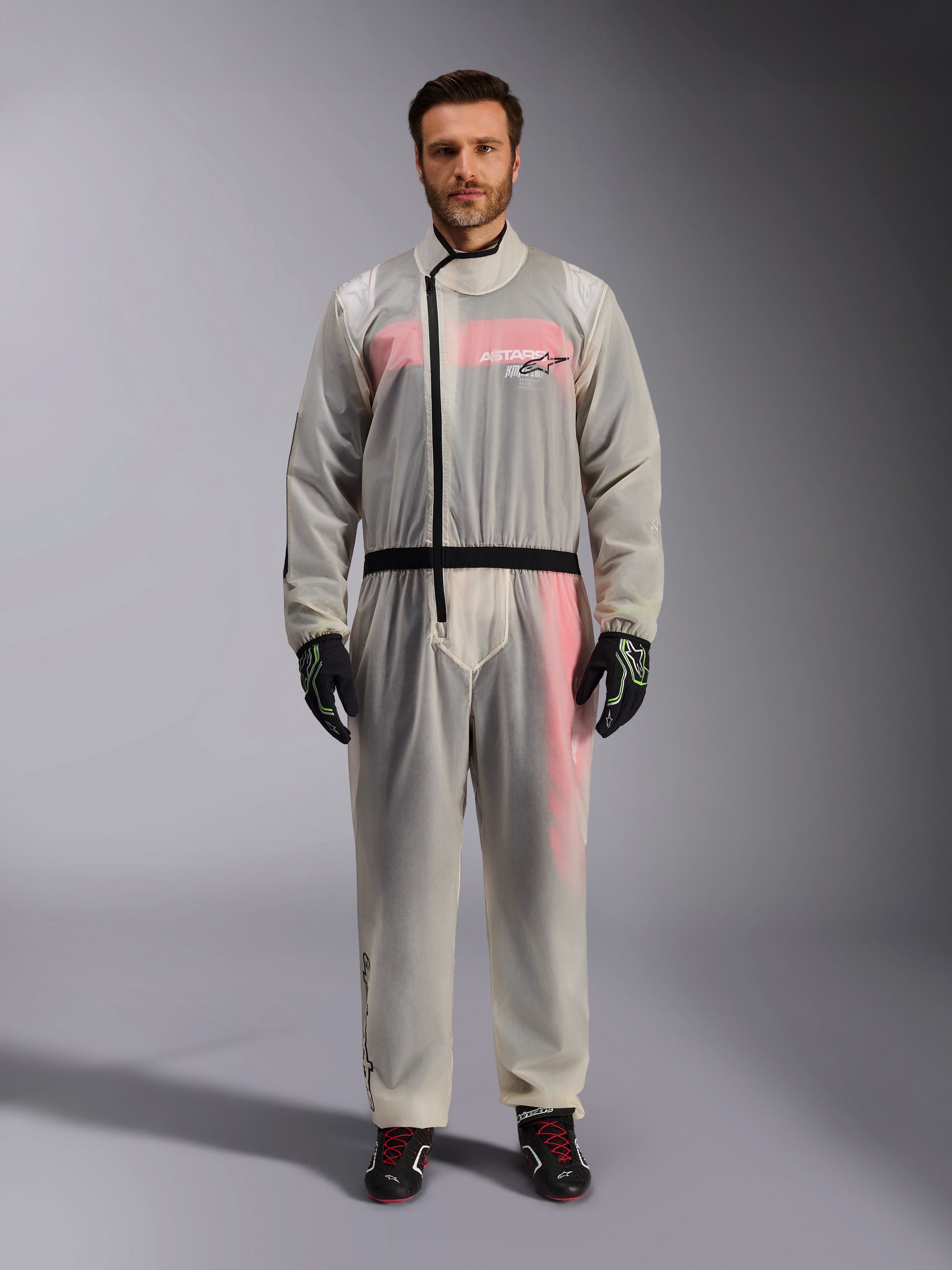 Alpinestars Kart Rain Suit, Clear, genomskinligt vattentätt regnställ som bärs av en modell, med en svart asymmetrisk dragkedja och logotyp på bröstet, röd och vit racingdräkt synlig under, matchat med svarta och neongröna handskar och svarta racingstövlar.