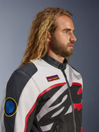 Man with long hair and beard wearing the Alpinestars Axton Jacket, Motorcycle Jacket, Black Off White and Bright Red, vintage racing-inspired design with branded patches and wrap-around logo, close-up profile view, urban contemporary style