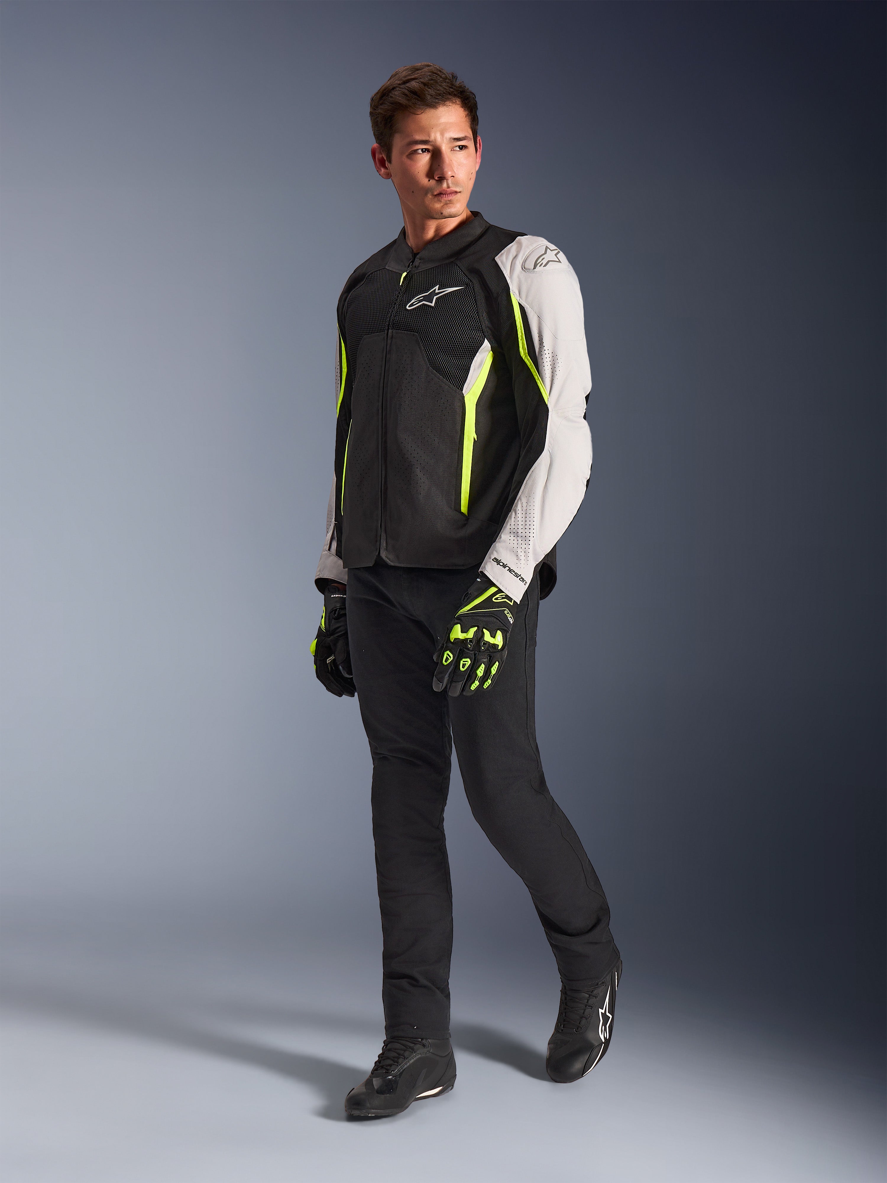 Alpinestars AST V3 Air Jacket, lätt motorcykeljacka i textil i svart, silver och Yellow Fluo, bärs av en modell med svarta byxor och matchande handskar, med meshpaneler och laserperforeringar för sportkörning i varmt väder.