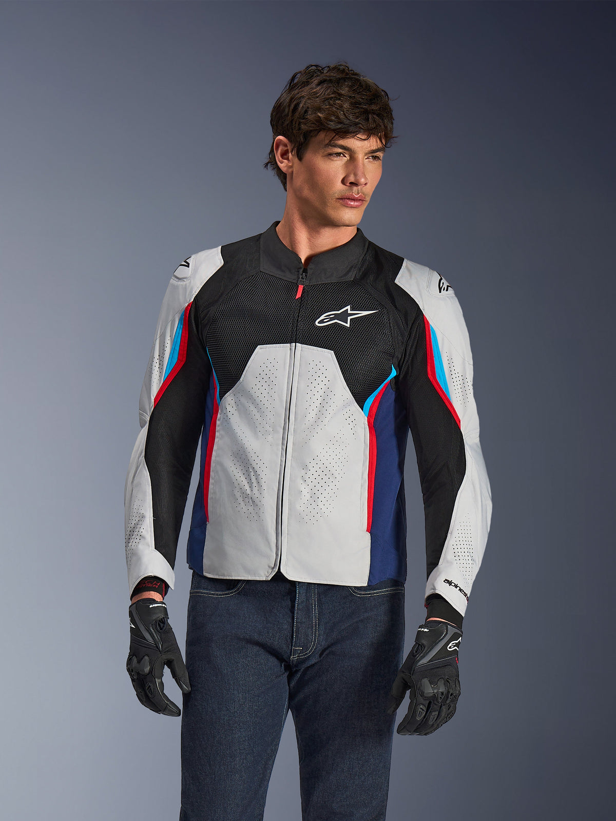 Alpinestars AST V3 Air Jacket i Silver Deep Blue Bright Red, bärs av en modell med mörka jeans och svarta handskar, med perforerade silverpaneler, svart mesh-ventilation och sportiga röda och blå accenter.