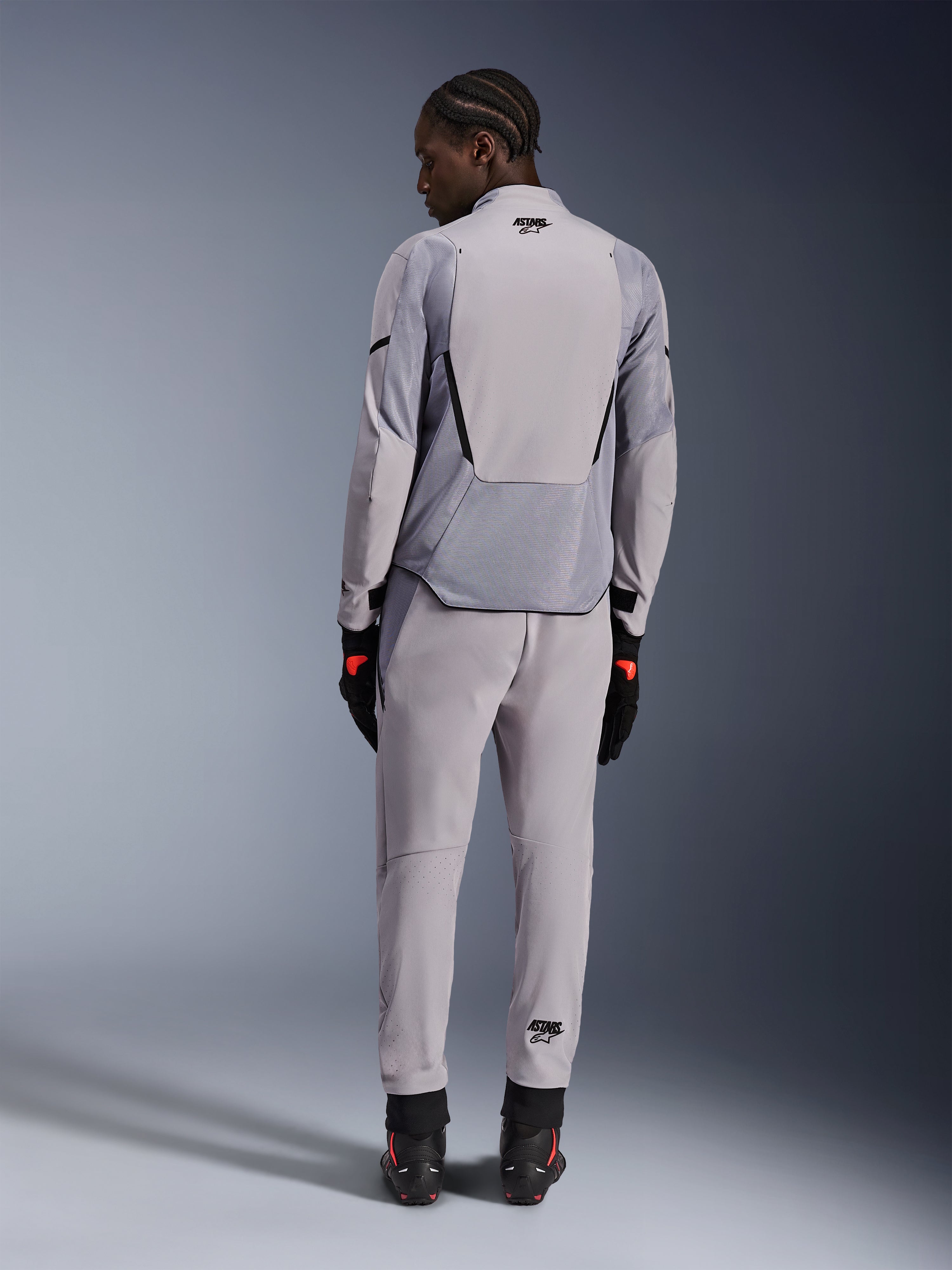 Alpinestars Aeroshell Airflow-jacka, Mid Gray Anthracite, sedd bakifrån på en modell, med tekniska meshpaneler och laserperforerade ventilationszoner, buren med matchande gråa byxor och svart motorcykelutrustning, sportskuren design för hög prestanda.