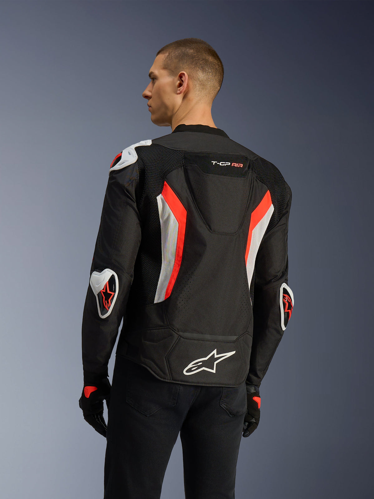 Alpinestars T-GP Air-jacka i Black Red Fluo, trekvartsvy bakifrån på en modell som framhäver den aerodynamiska puckeln, externa Racing GP DFS-skydd på axlar och armbågar, laserperforerade meshpaneler samt vita och fluorescerande röda detaljer, sportskuren textiljacka för motorcykel