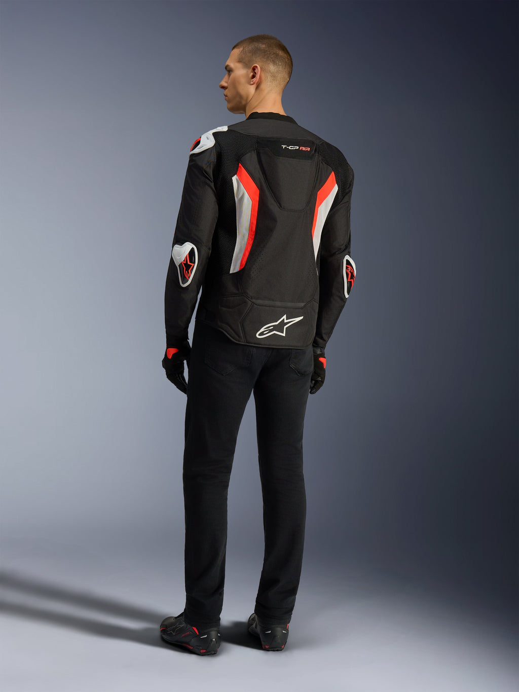 Alpinestars T-GP Air Jacket, svart röd fluo motorcykeljacka, sedd bakifrån på en modell, med ventilationspaneler i mesh, aerodynamisk puckel och vita externa skydd på armbågar och axlar, bärs med svarta byxor och körskor.