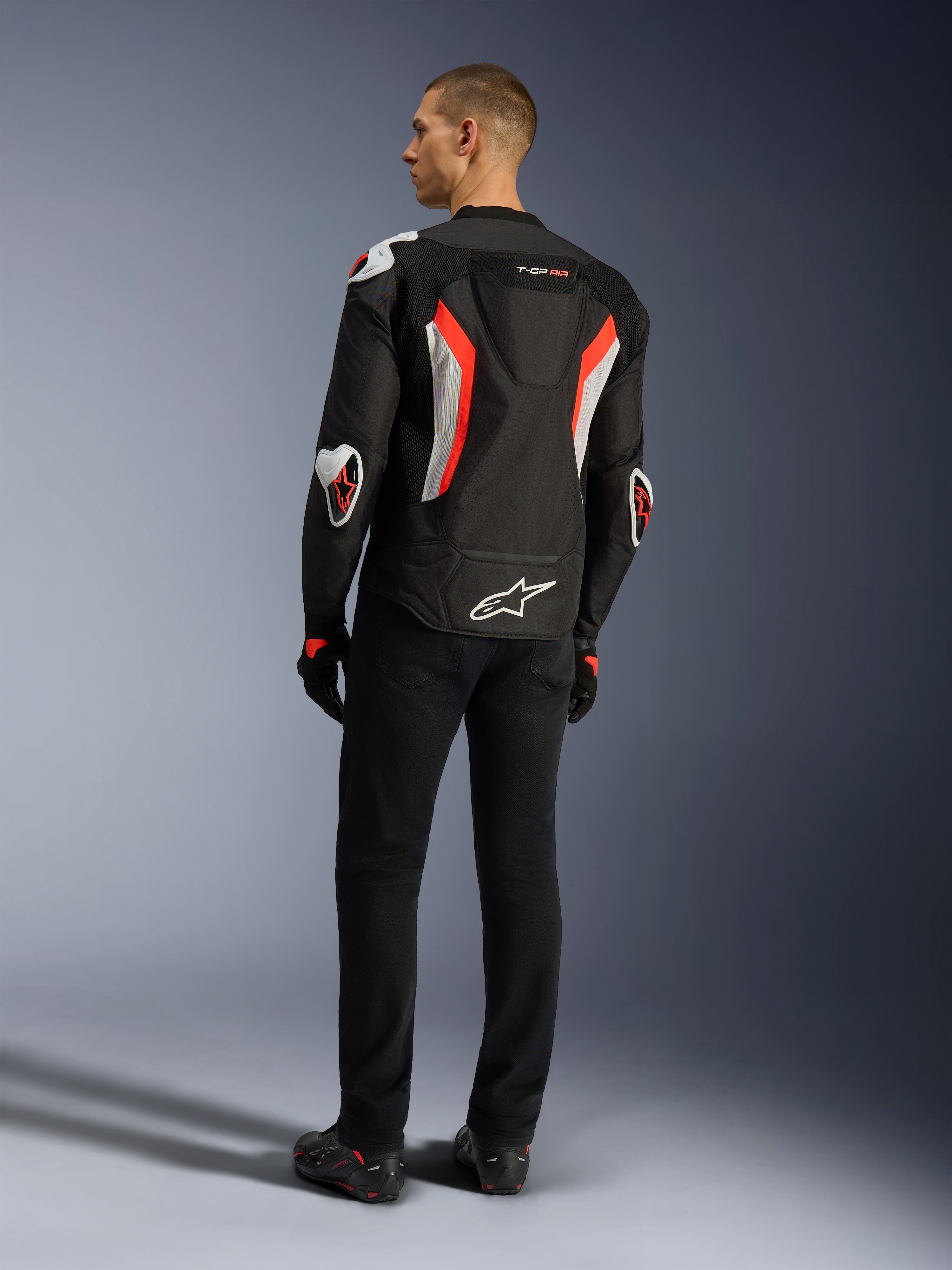 Alpinestars T-GP Air Jacket, svart röd fluo motorcykeljacka, sedd bakifrån på en modell, med ventilationspaneler i mesh, aerodynamisk puckel och vita externa skydd på armbågar och axlar, bärs med svarta byxor och körskor.