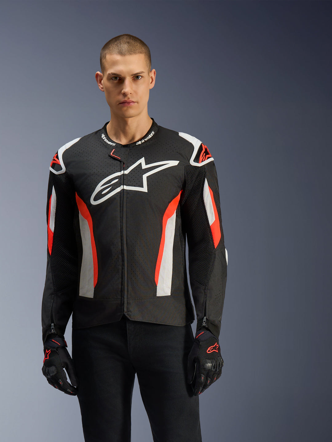 Manlig modell som bär Alpinestars T-GP Air-jacka, svart med röd fluo och vita detaljer, stor Alpinestars-logotyp på bröstet, med laserperforerade paneler och axelskydd, tillsammans med svarta körhandskar, sportigt skuren motorcykelutrustning för varmt väder