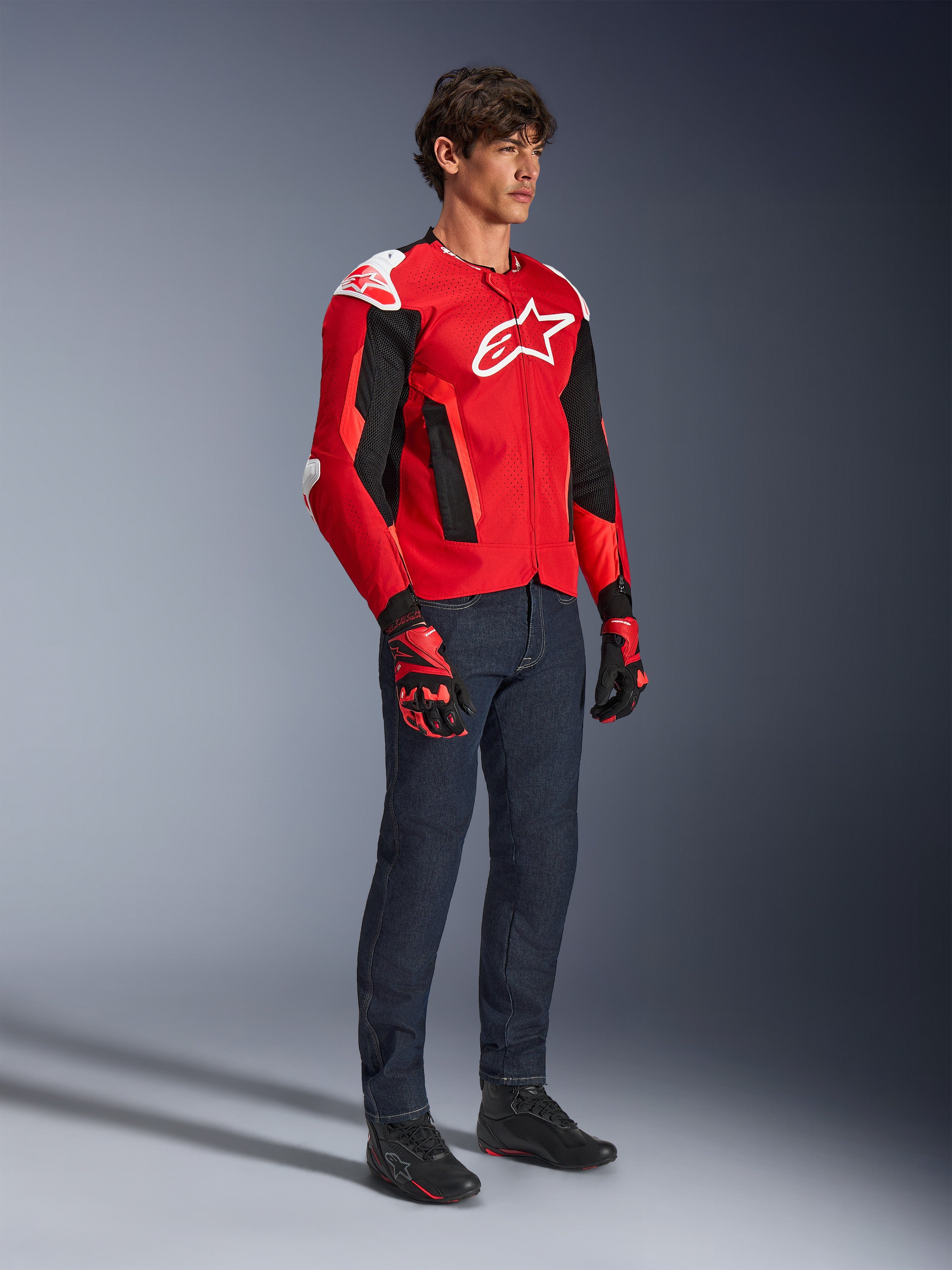 Alpinestars T-GP Air Jacket, Sport Motorcycle Jacket, Bright Red and Black with white accents, worn by a model with dark blue jeans and red racing gloves, featuring high-flow mesh panels and white shoulder sliders, aerodynamic sport fit for warm weather riding.