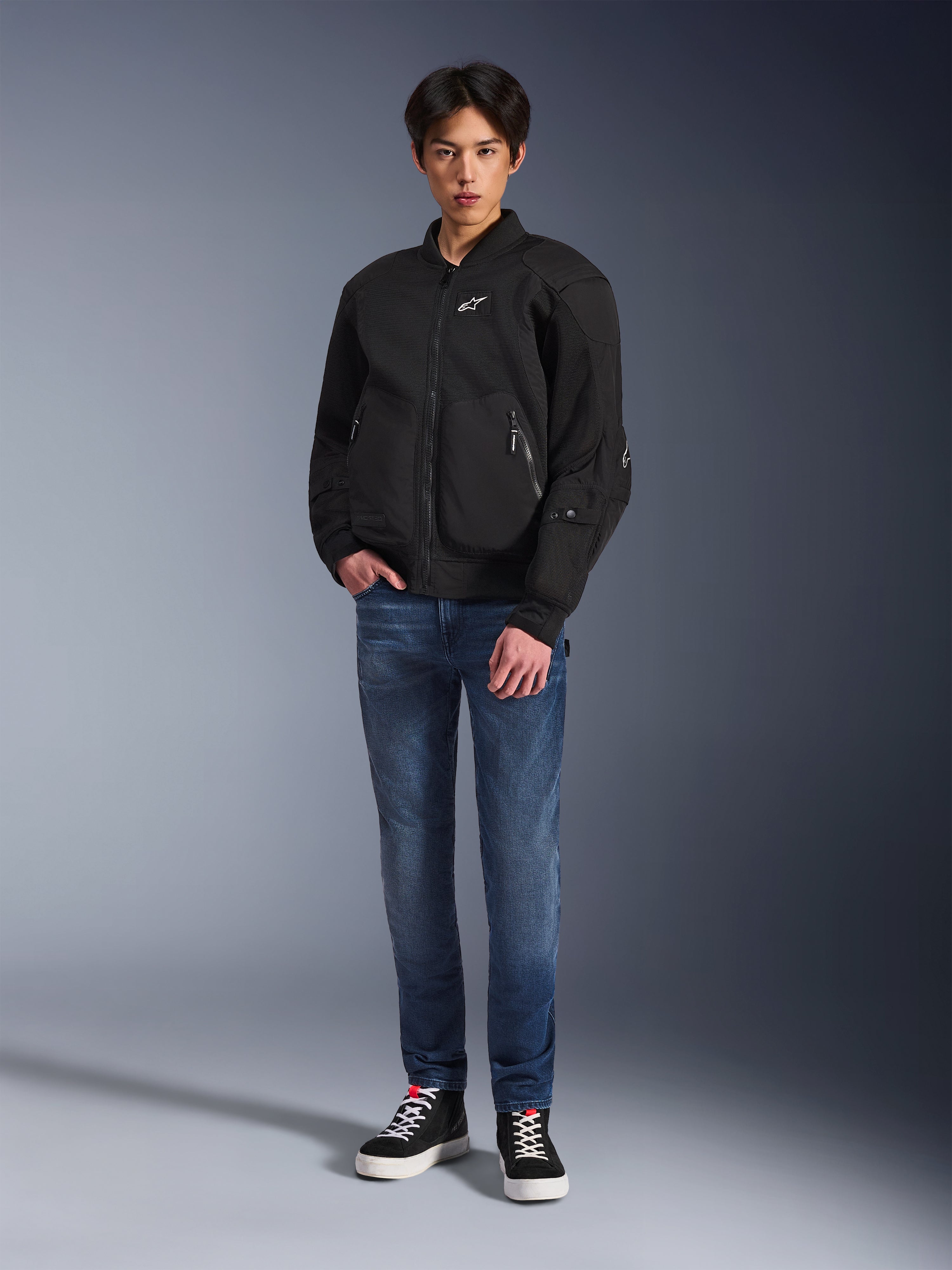 Model wearing the Alpinestars Flight Air Jacket in Black, an urban bomber-style motorcycle jacket with 3D mesh panels and a front zipper, paired with blue jeans and black sneakers, contemporary street-inspired riding apparel.
