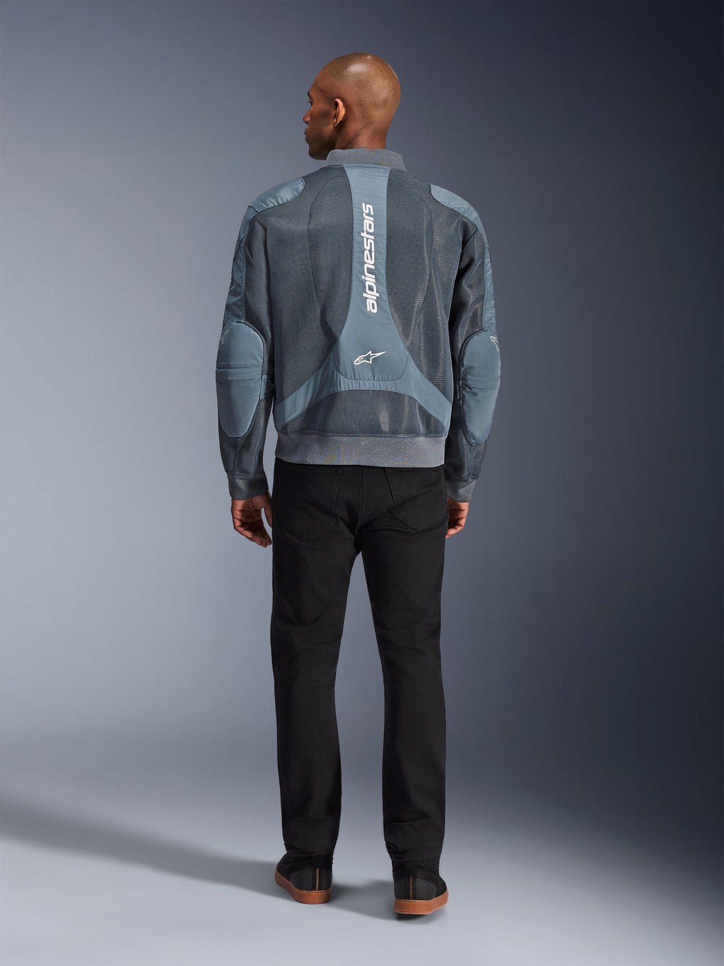 Alpinestars Flight Air-jacka i Storm Gray, baksidesvy av manlig modell i en motorcykeljacka i urban stil med 3D-meshpaneler och vertikal Alpinestars-logotyp, matchad med svarta byxor för en modern pendlarlook