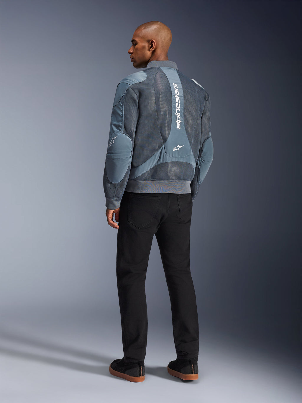 Alpinestars Flight Air-jacka, stormgrå motorcykeljacka, sedd bakifrån, med ventilationspaneler i 3D-mesh och vertikal vit Alpinestars-logotyp, bärs med svarta byxor och svarta sneakers, urban pendlingsstil