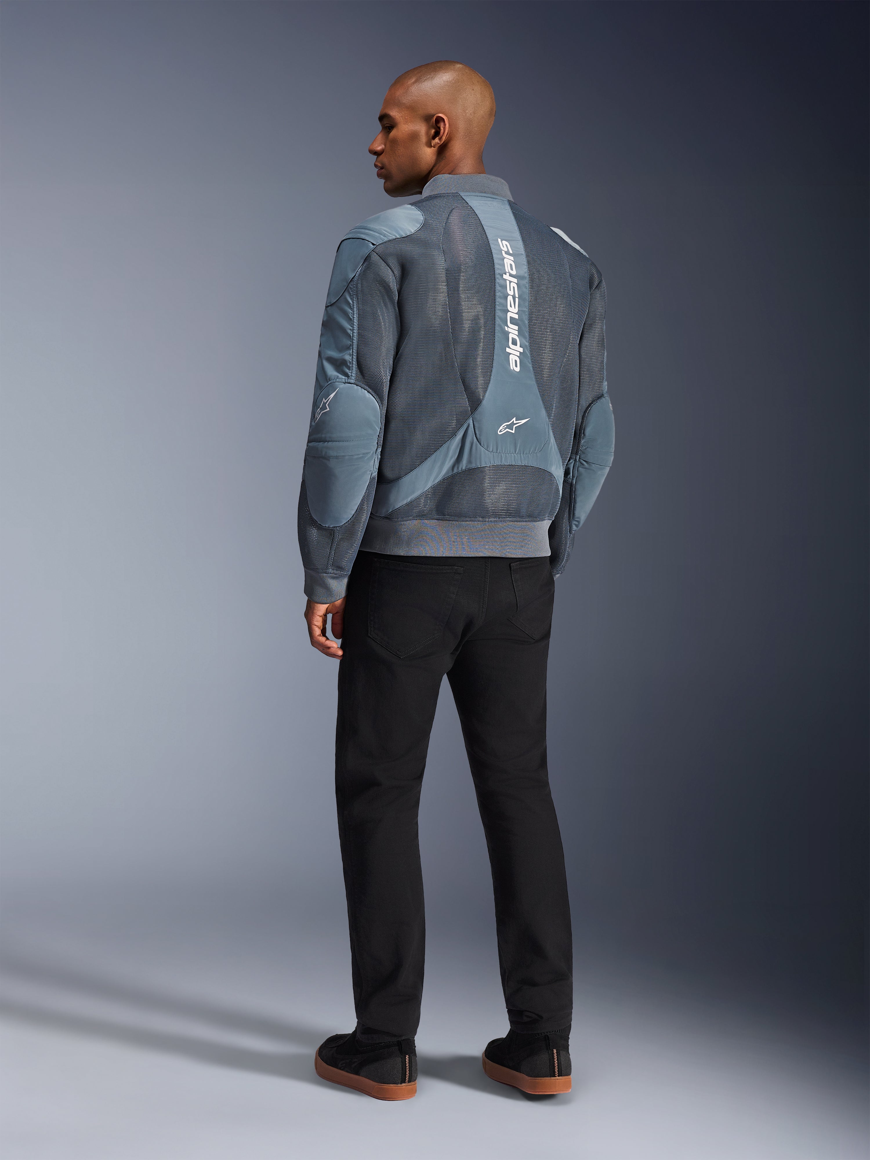 Alpinestars Flight Air-jacka, stormgrå motorcykeljacka, sedd bakifrån, med ventilationspaneler i 3D-mesh och vertikal vit Alpinestars-logotyp, bärs med svarta byxor och svarta sneakers, urban pendlingsstil