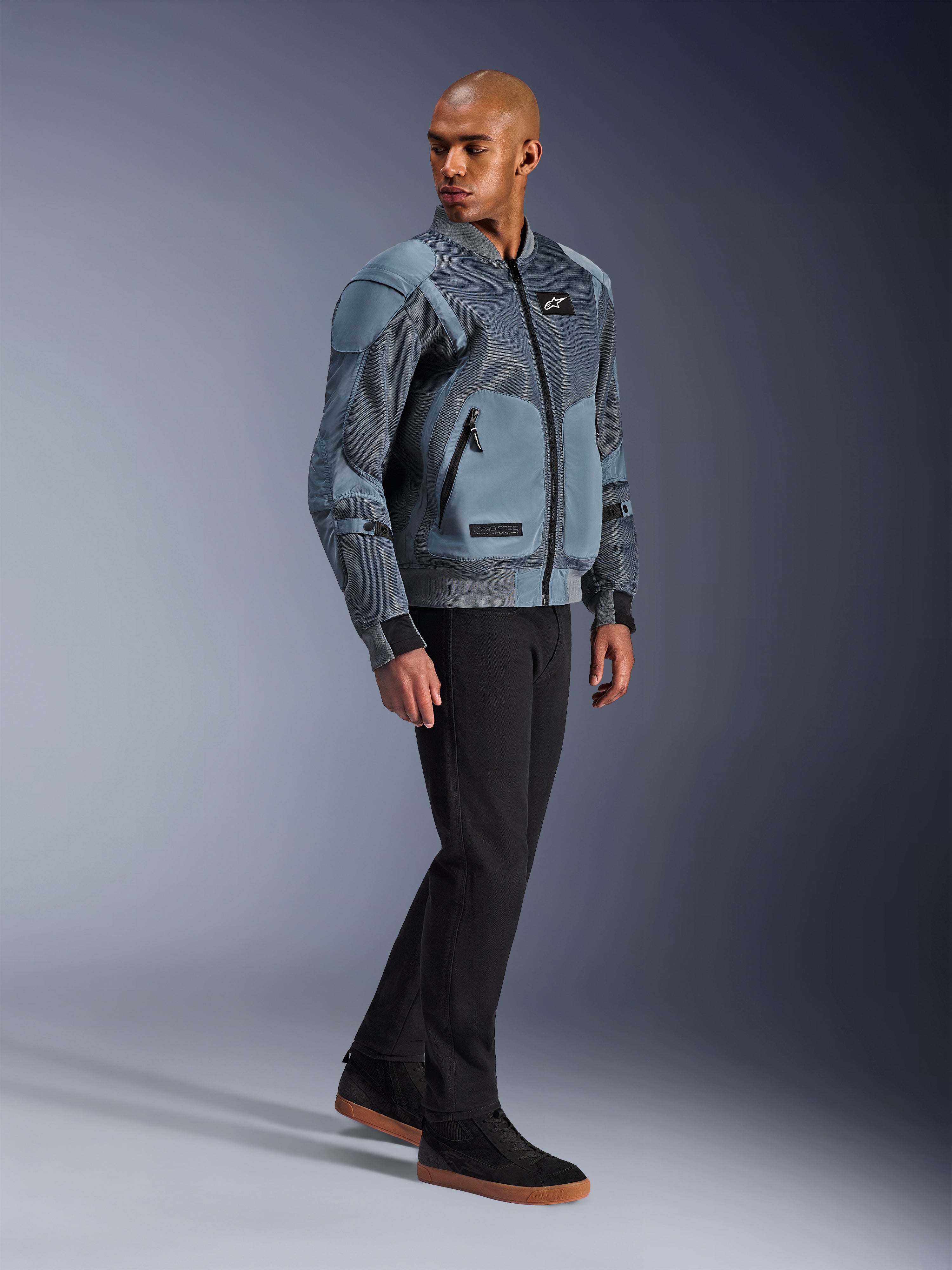 Alpinestars Flight Air-jacka, Storm Gray, urban bomber-inspirerad motorcykeljacka, bärs av en manlig modell med svarta byxor och sneakers, utrustad med utökade ventilationspaneler i 3D-mesh och externa skyddsfickor, modern streetwear-skyddsutrustning för körning i varmt väder