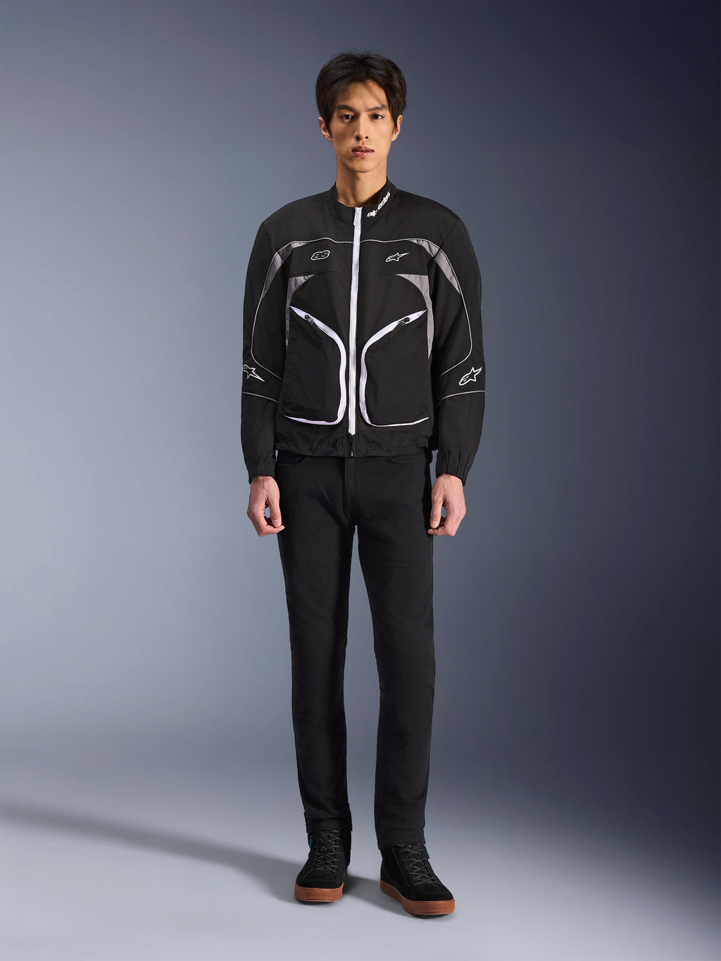Alpinestars Unite Jacket, Motorcycle Jacket, Black and Dark Gray, worn by a model with black pants and black boots, featuring white contrast zippers, reflective piping, and minimalist urban design