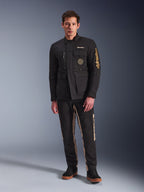 Male model wearing the Alpinestars Dice Jacket, Black urban motorcycle jacket with vintage field jacket styling, four functional front pockets, Alpinestars branding on the sleeve and chest, paired with black and tan pants, modern urban riding gear