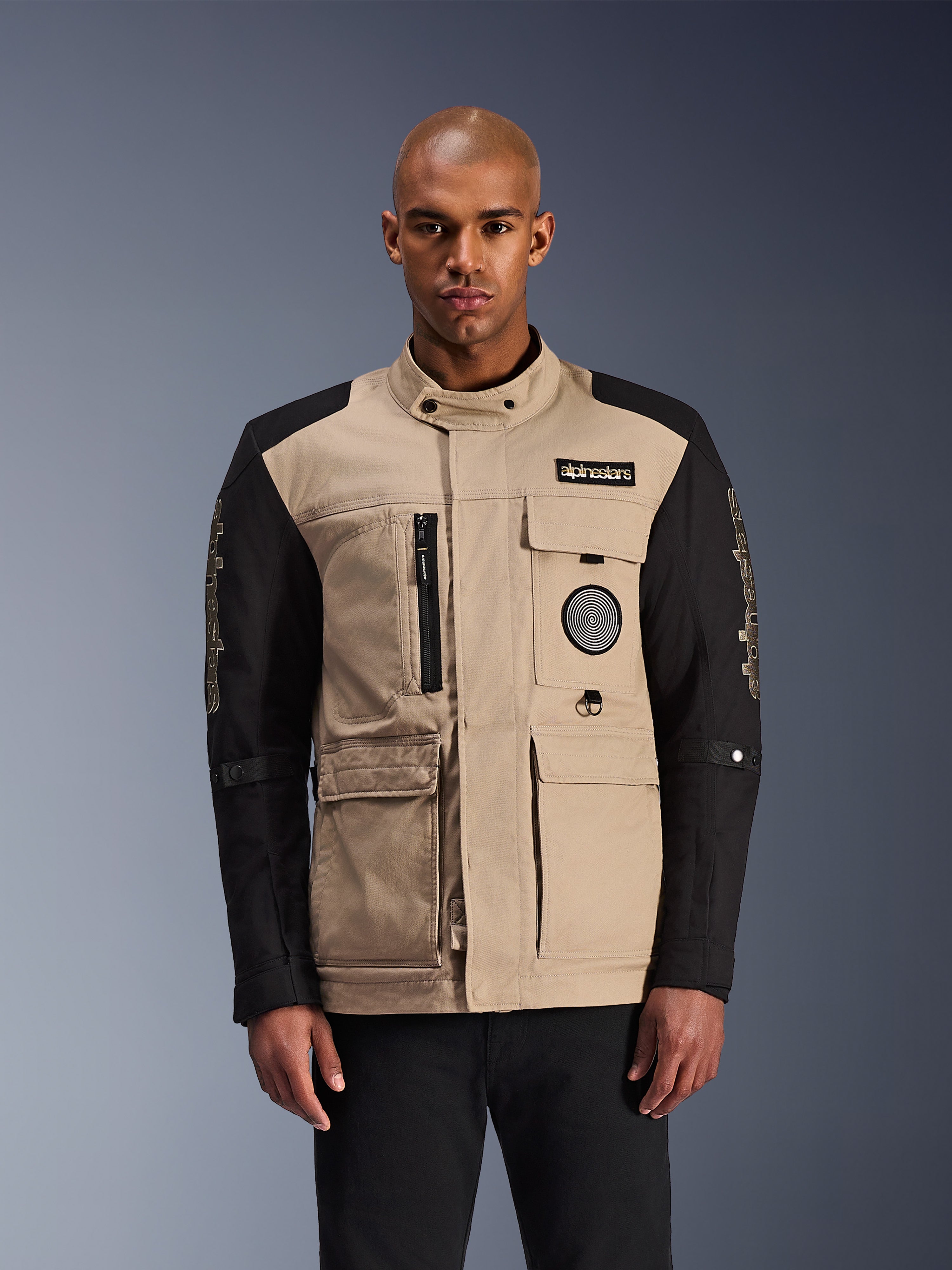 Alpinestars Dice Jacket, urban motorcykeljacka, beige och svart, robust arbetsklädesestetik med flera praktiska fickor och bröstmärken, hög krage med knappstängning, tydlig logotyp på ärmarna, bärs av en manlig modell