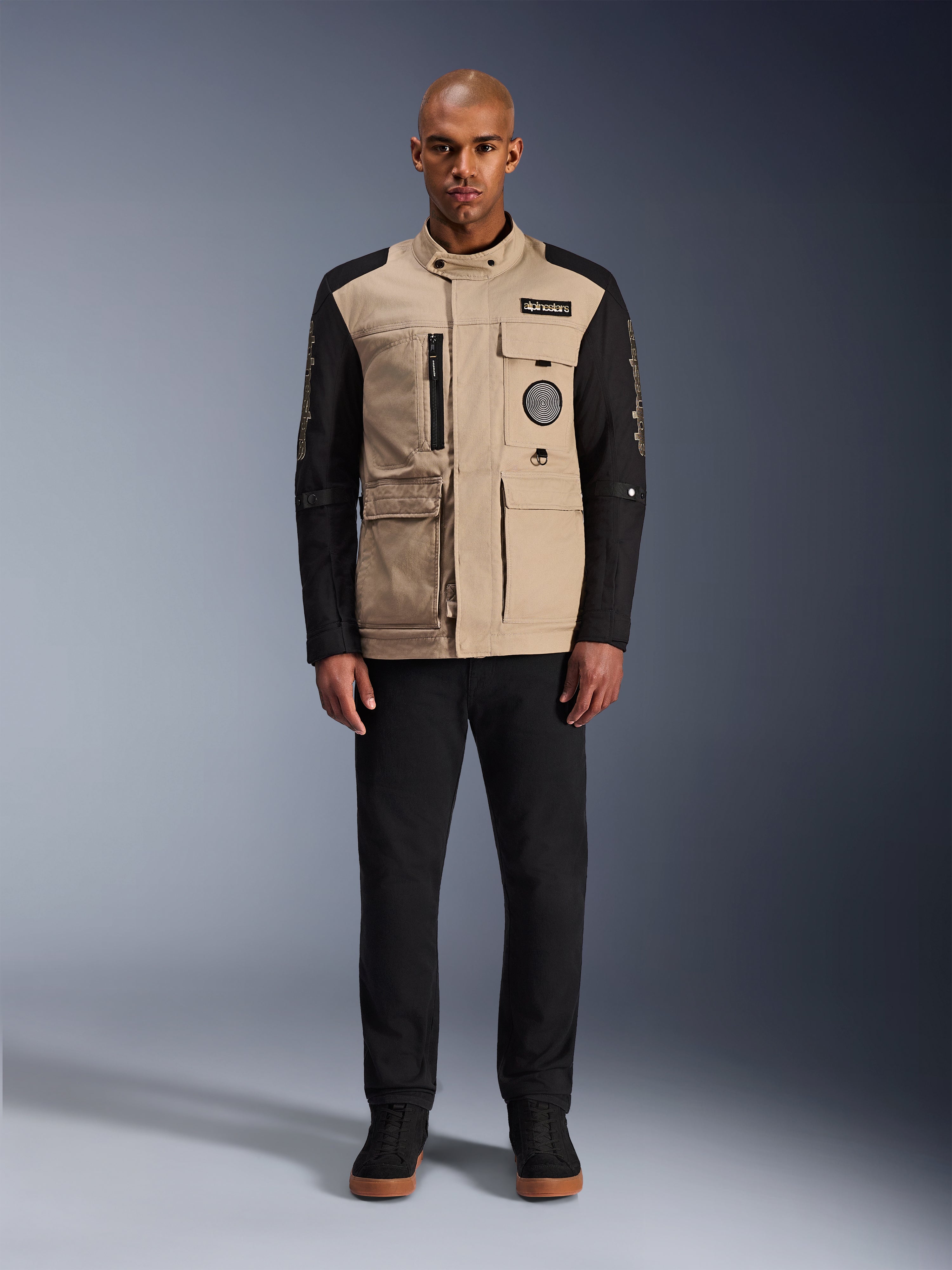 Alpinestars Dice Jacket, urban motorcykeljacka, aluminium-beige huvuddel med svarta ärmar, arbetsklädesinspirerad silhuett med fyra praktiska framfickor, bärs med svarta byxor och svarta high-top sneakers, robust körutrustning redo för gatan