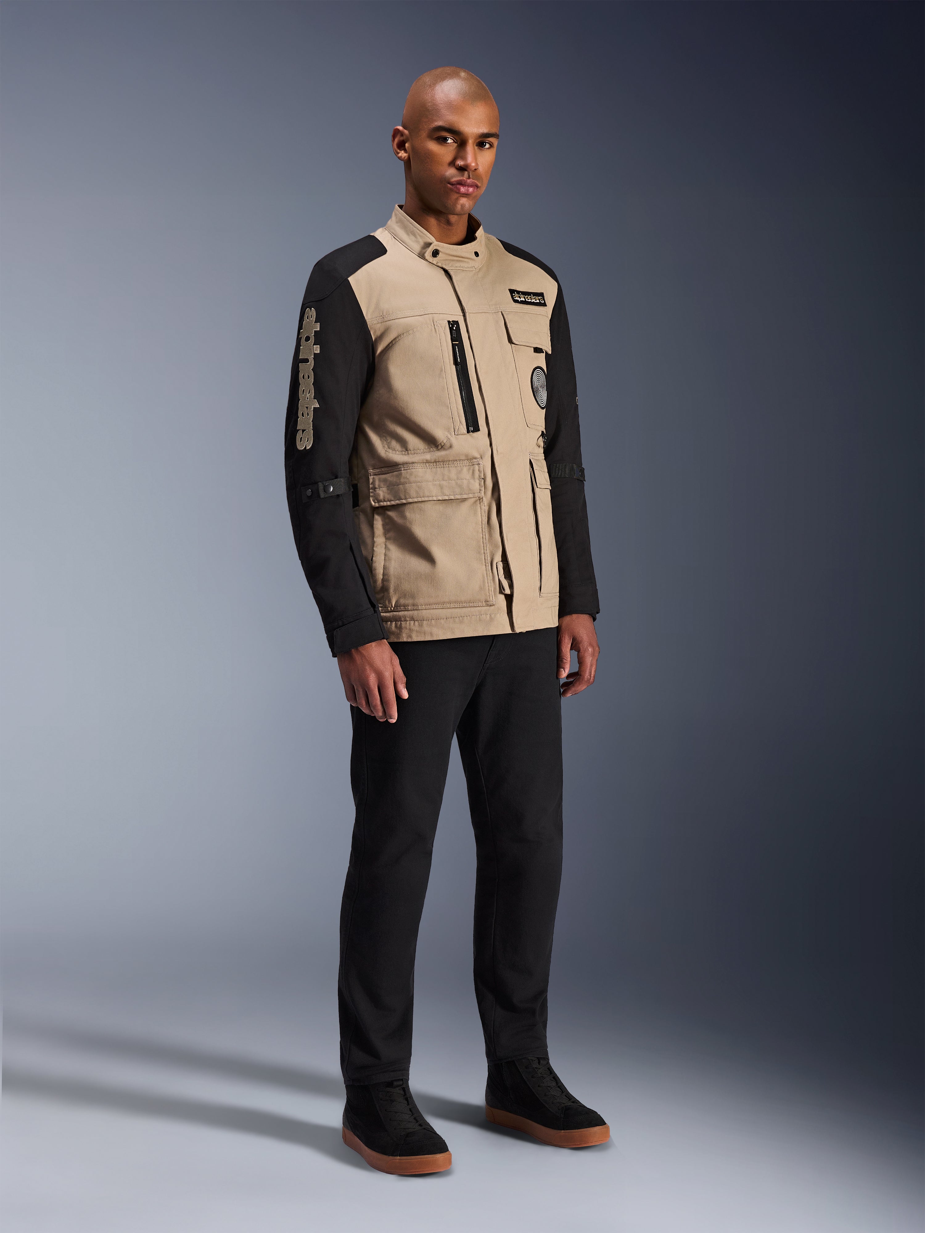 Alpinestars Dice Jacket, urban motorcykeljacka, Aluminum Black och khaki, tvåtonad design med svarta ärmar och khakifärgad torso, bärs av en modell med svarta byxor och svarta sneakers, arbetsklädesinspirerad estetik med praktiska fickor och broderad logotyp på ärmen.