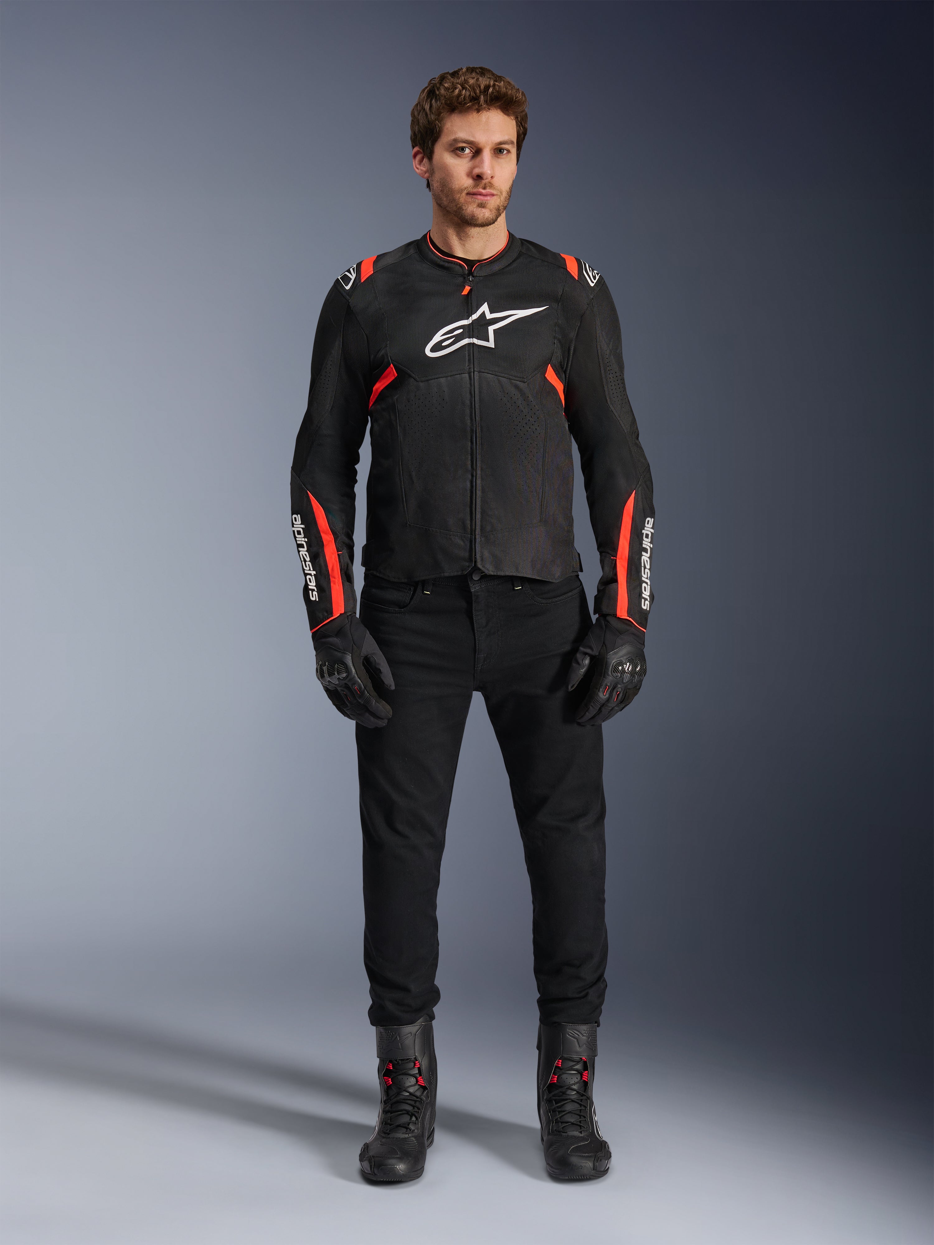 Male model wearing Alpinestars T-SPS Air v2 Jacket, Black textile motorcycle jacket with Red Fluo accents, white logo on chest, worn with black pants, gloves and boots, urban sport riding style