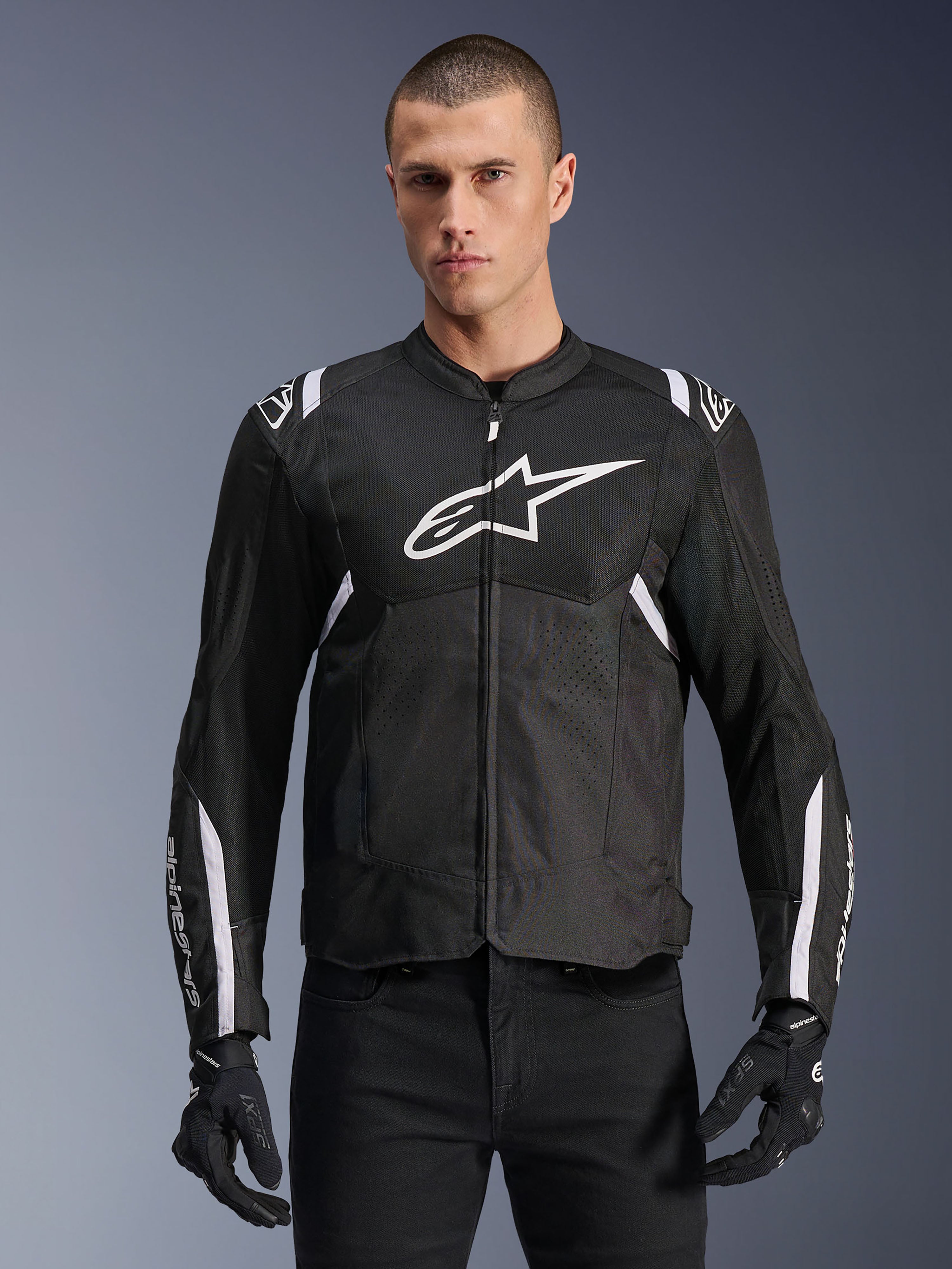 Alpinestars T-SPS Air V2 jacka, textil motorcykeljacka, svart med vita detaljer, stor vit bröstlogotyp, perforerad mesh-design för ventilation, bärs med svarta byxor och handskar