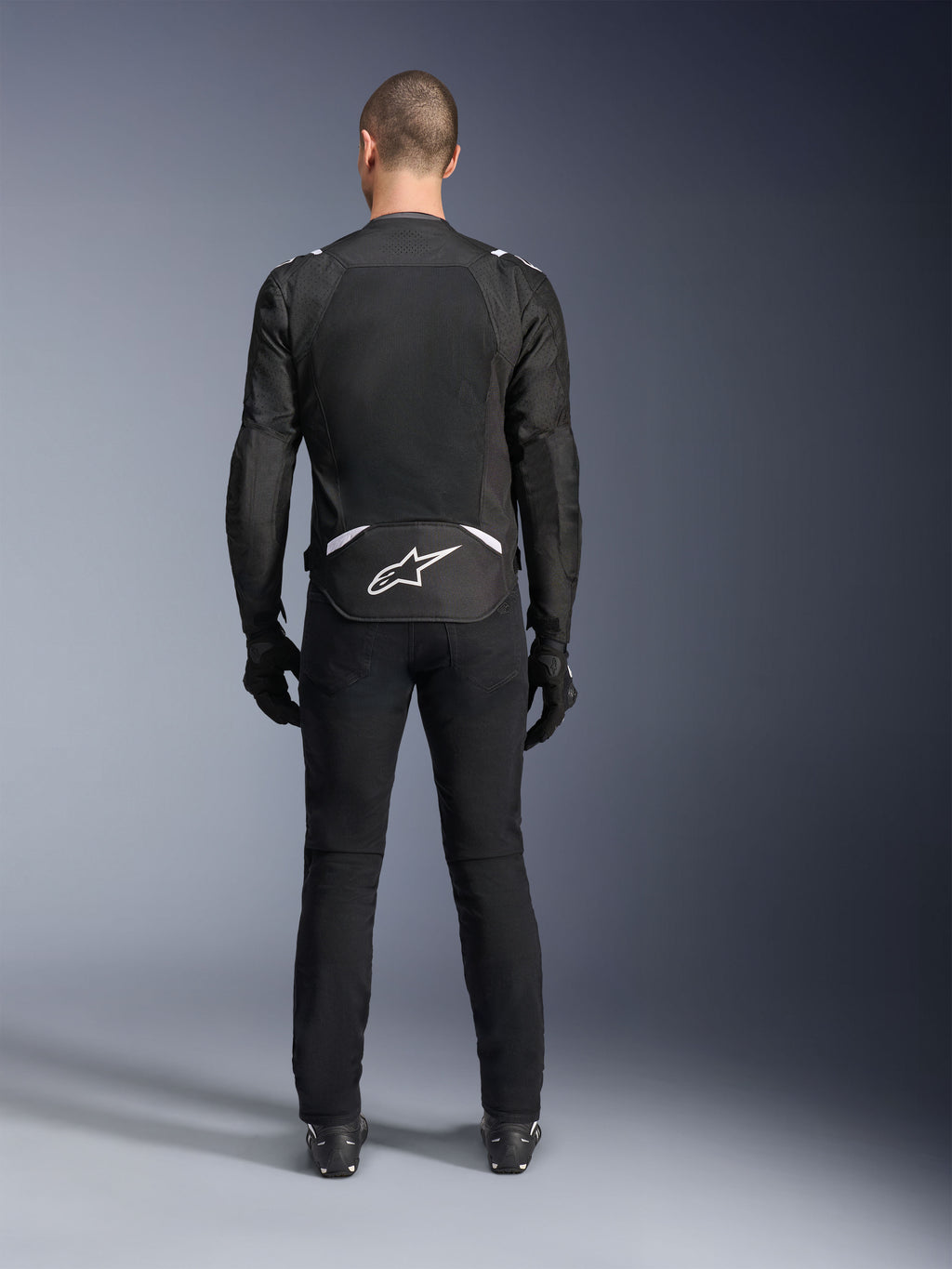 Alpinestars T-Sps Air V2-jacka, sedd bakifrån, svart och vit motorcykeljacka i textil, med en stor meshpanel på ryggen för maximalt luftflöde och vit Astars-logotyp på nedre ryggen, bärs med svarta mc-byxor och handskar
