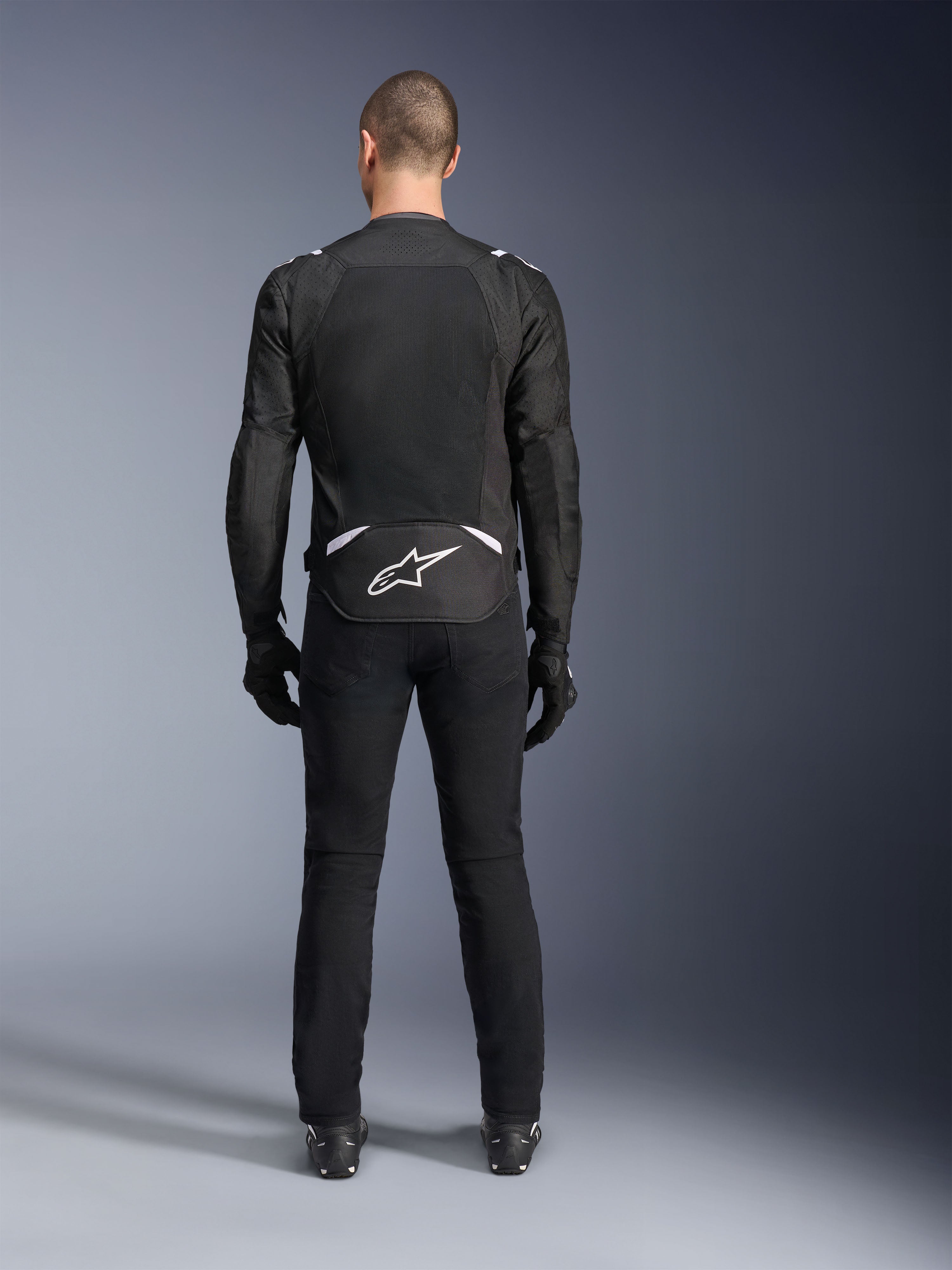 Alpinestars T-Sps Air V2-jacka, sedd bakifrån, svart och vit motorcykeljacka i textil, med en stor meshpanel på ryggen för maximalt luftflöde och vit Astars-logotyp på nedre ryggen, bärs med svarta mc-byxor och handskar