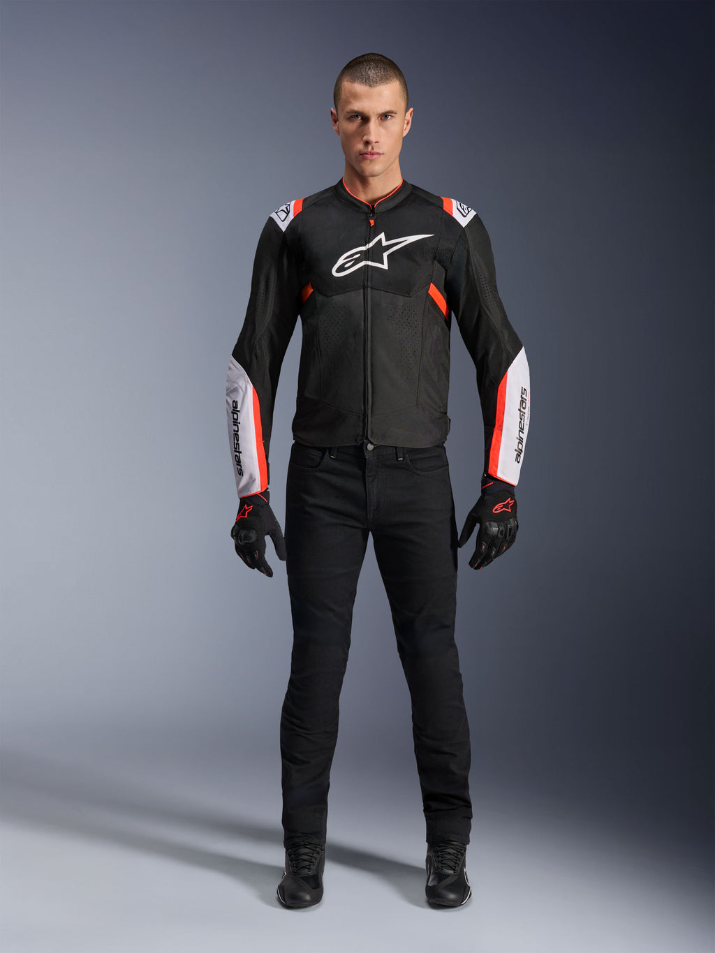 Manlig modell som bär Alpinestars T-Sps Air V2-jacka i Black White Red Fluo, med stora tech-mesh-paneler och framträdande vit logotyp på bröstet och ärmarna, matchad med svarta tekniska körbyxor och handskar, urban sport-motorcykelutrustning