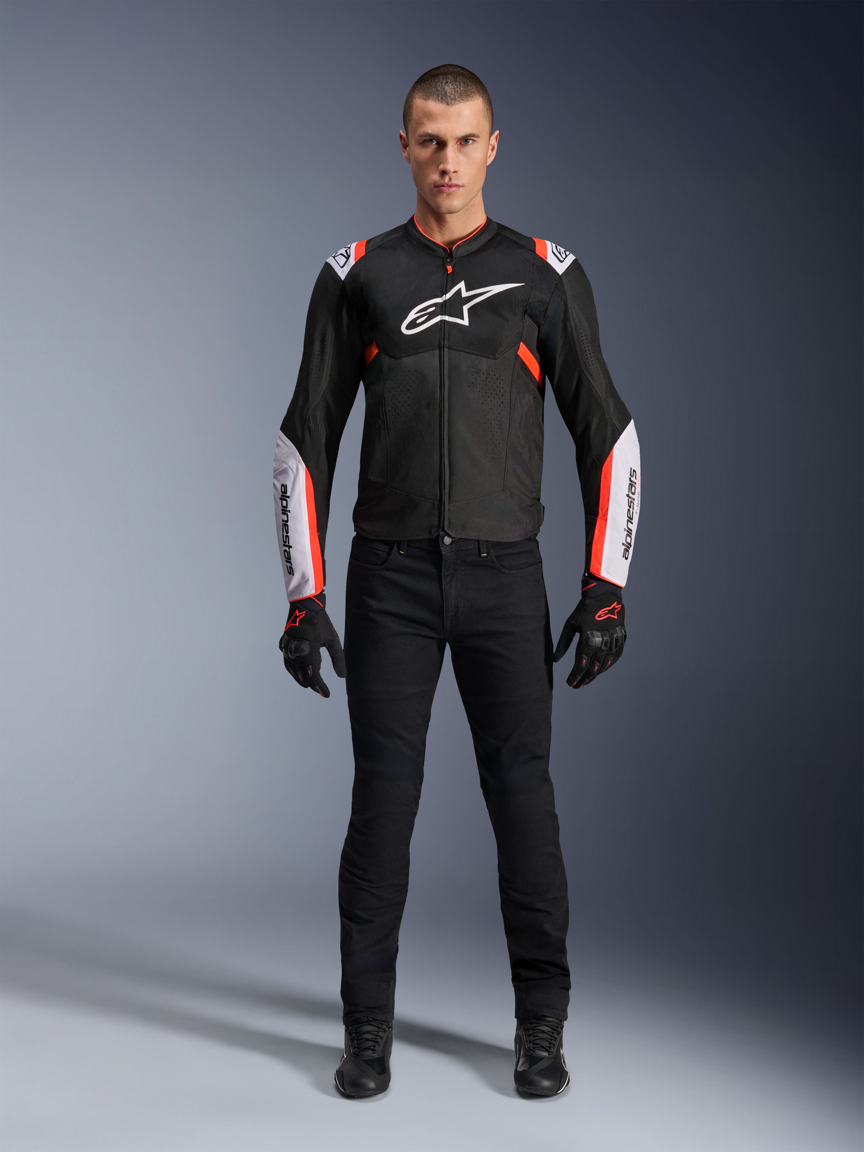 Manlig modell som bär Alpinestars T-Sps Air V2-jacka i Black White Red Fluo, med stora tech-mesh-paneler och framträdande vit logotyp på bröstet och ärmarna, matchad med svarta tekniska körbyxor och handskar, urban sport-motorcykelutrustning