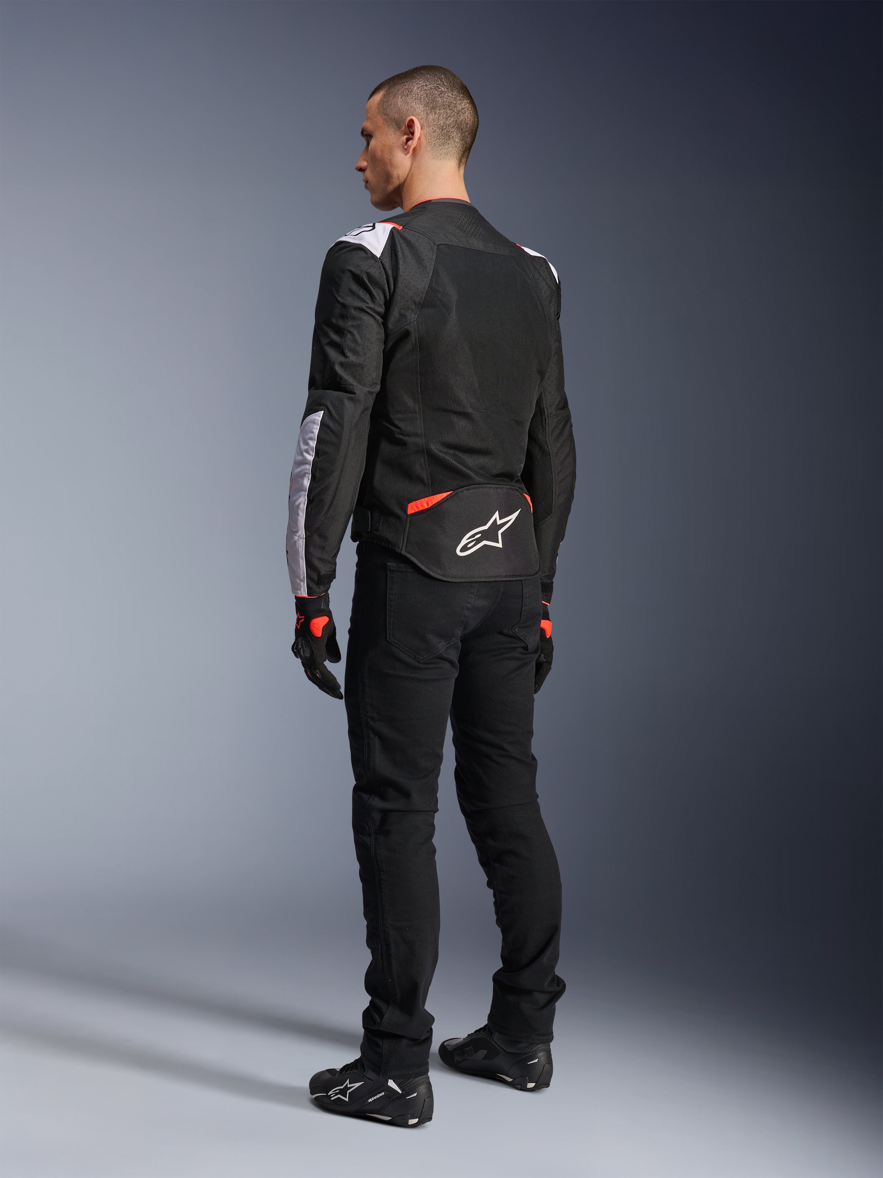 Alpinestars T-Sps Air V2-jacka, motorcykeljacka i textil, svart, vit och röd fluo, vy bakifrån på modell, meshpaneler för ventilation, vit Astars-logotyp på nedre delen av ryggen, stylad med svarta körbyxor och handskar