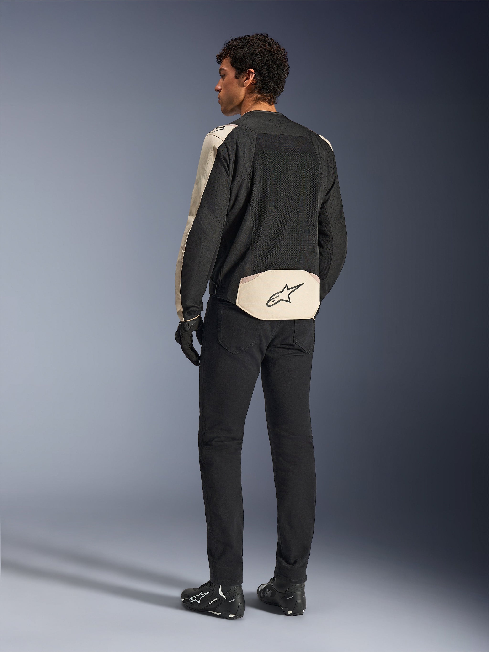 Alpinestars T-SPS Air V2-jacka, textiljacka för motorcykel, Black Mastic Aluminum, baksida buren av modell, med omfattande tech mesh-paneler för luftflöde, ljusbeige detaljer på ärmar och ländrygg med Alpinestars-logotypen, matchad med svarta byxor och stövlar.