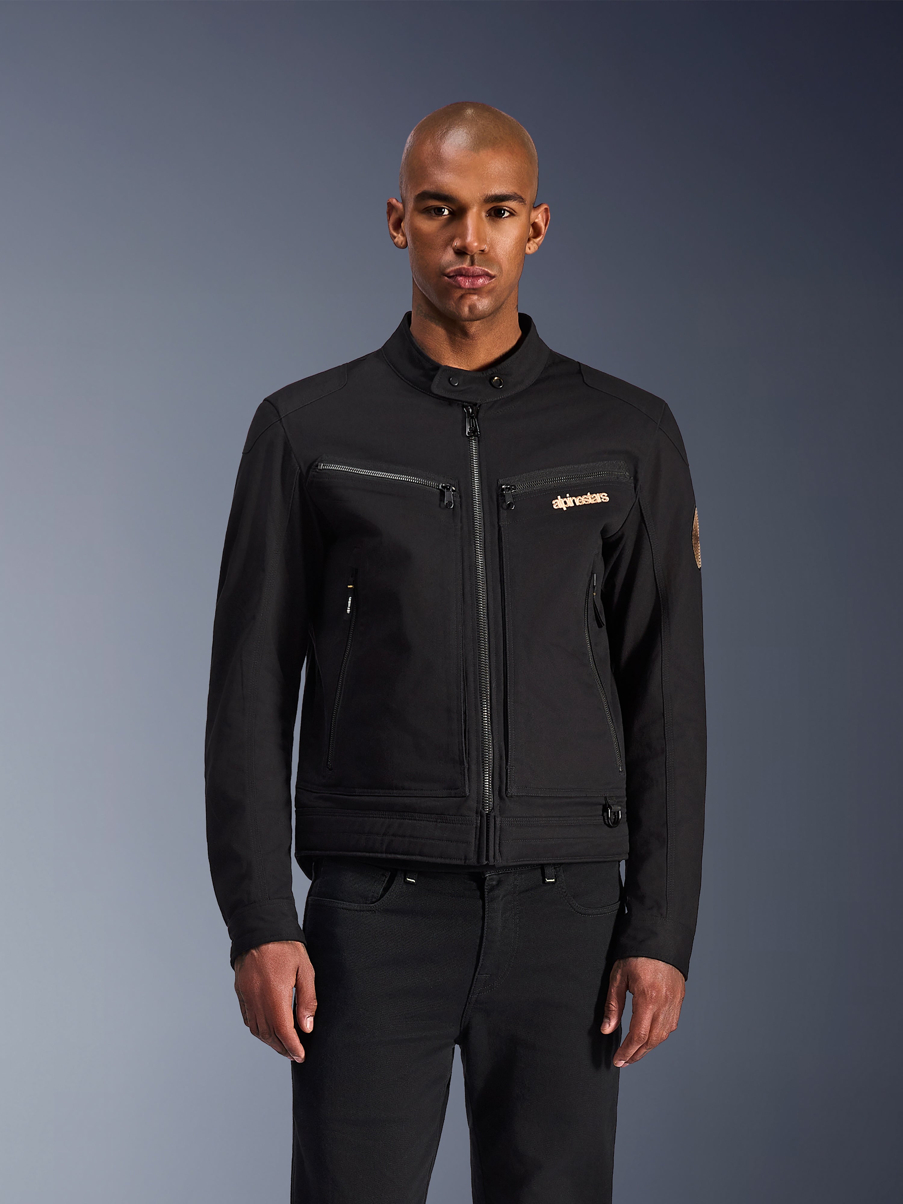 Alpinestars Bruiser-jacka, svart urban motorcykeljacka, framifrån på en modell, med klassisk arbetsklädesestetik, bröstfickor med dragkedja, en knäppt krage och Alpinestars-logotyp, designad för stadsförare