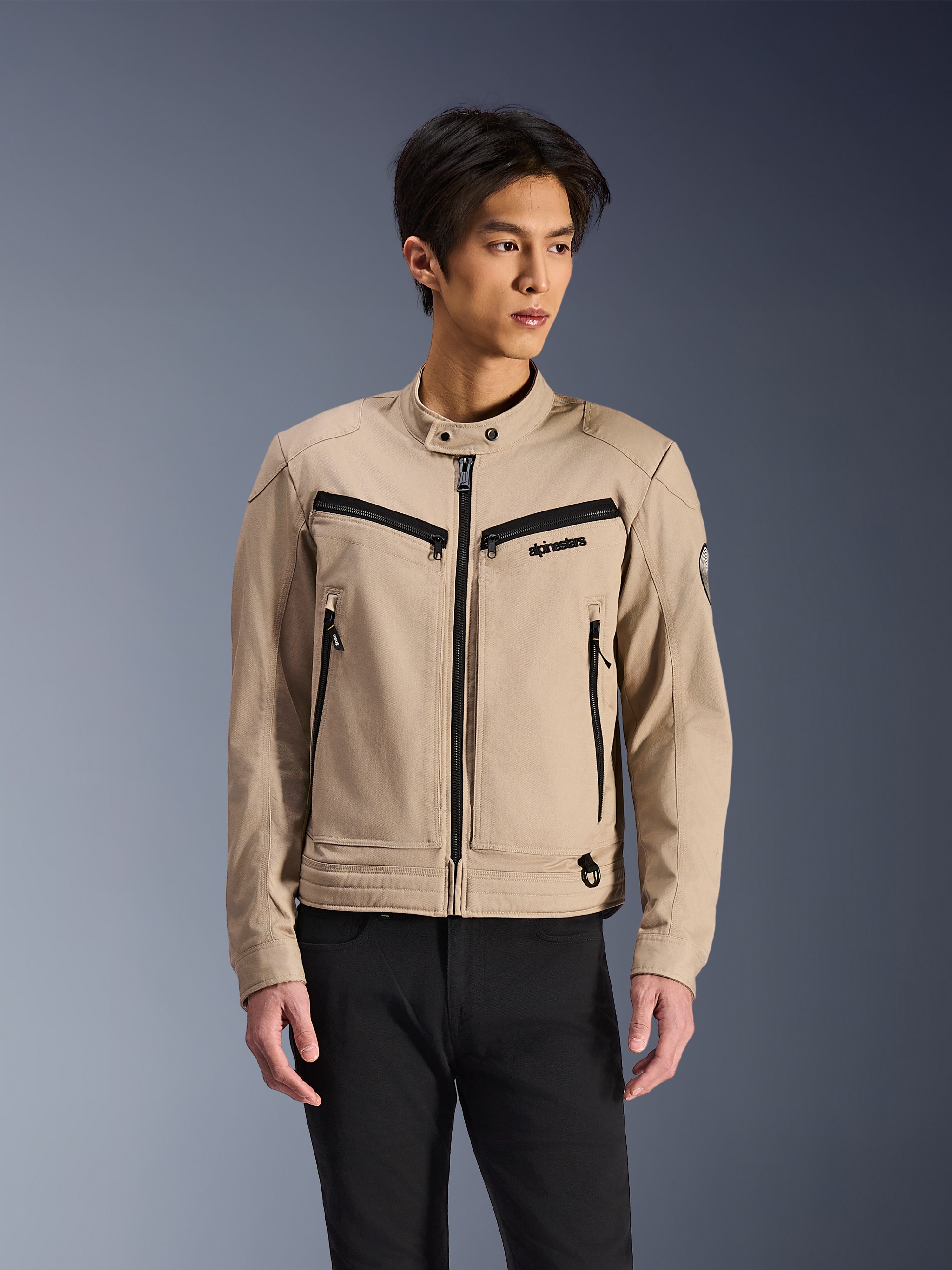 Alpinestars Bruiser-jacka, beige motorcykeljacka buren av manlig modell, med svarta dragkedjedetaljer på bröst och sidofickor, hög krage med tryckknapp, arbetsklädesinspirerad urban mc-stil, kombinerad med svarta byxor