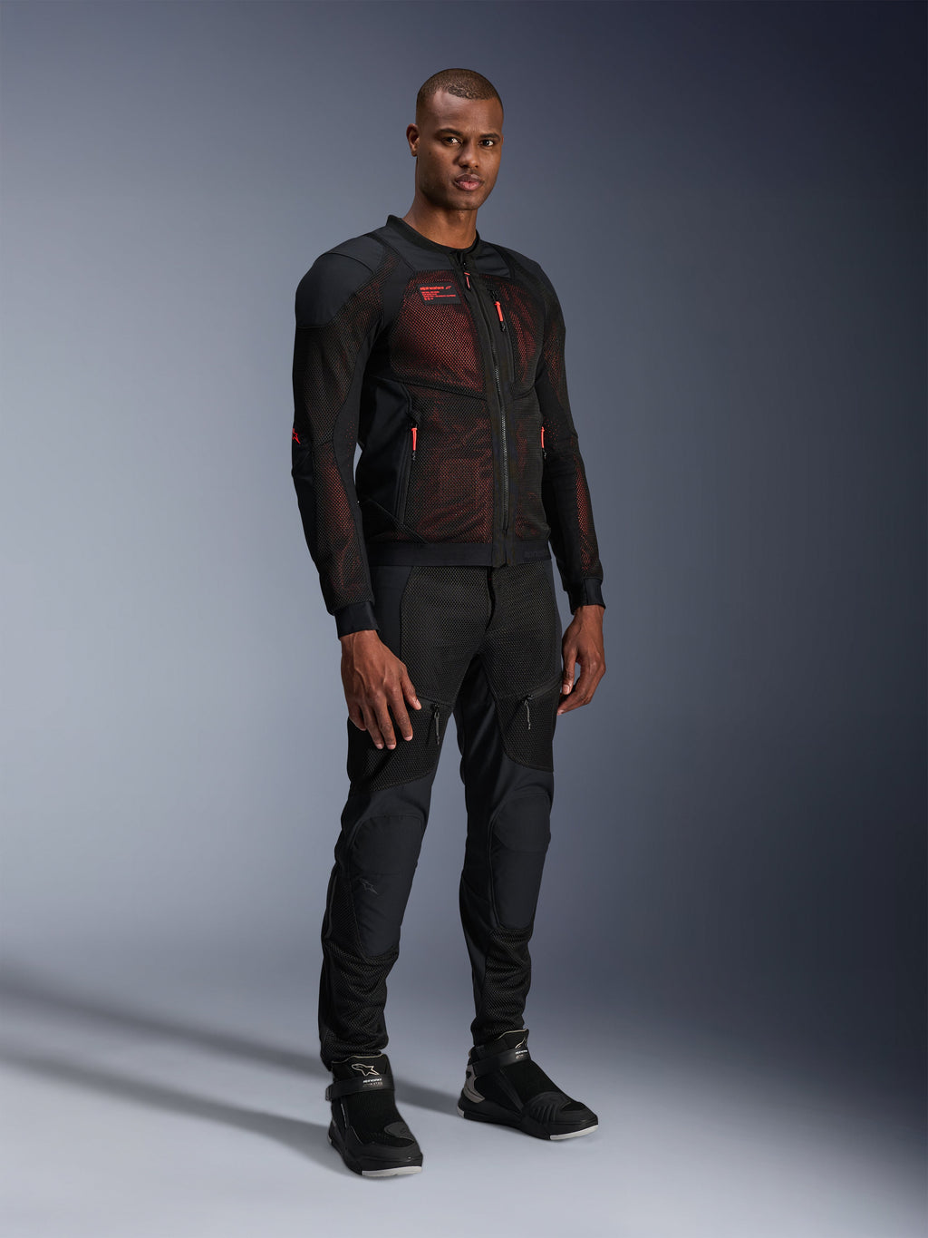 Alpinestars Troop-Air Pro-jacka i Black Red Fluo, teknisk motorcykeljacka i mesh med integrerade skydd, bärs av en manlig modell med matchande svarta skyddsförstärkta byxor och höga boots, studiofotografi i helfigur.