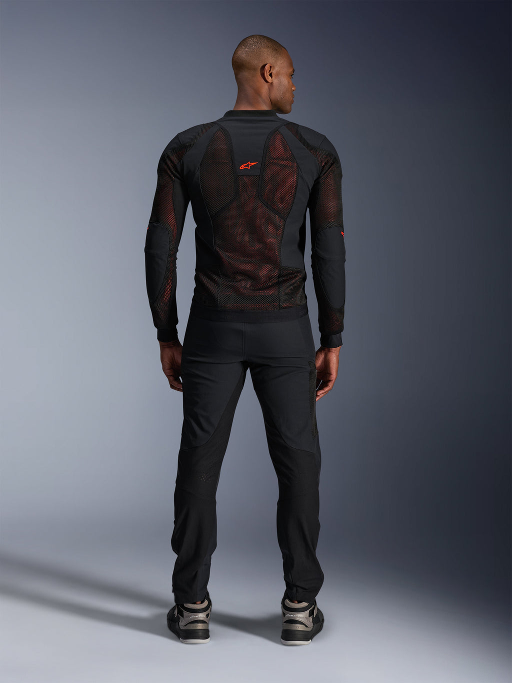 Alpinestars Troop-Air Pro-jacka i Black Red Fluo, vy bakifrån på modell, teknisk meshjacka med skydd och ventilerande paneler, röd logotyp på övre delen av ryggen, bärs med svarta byxor och sneakers, urban motorcykelutrustning
