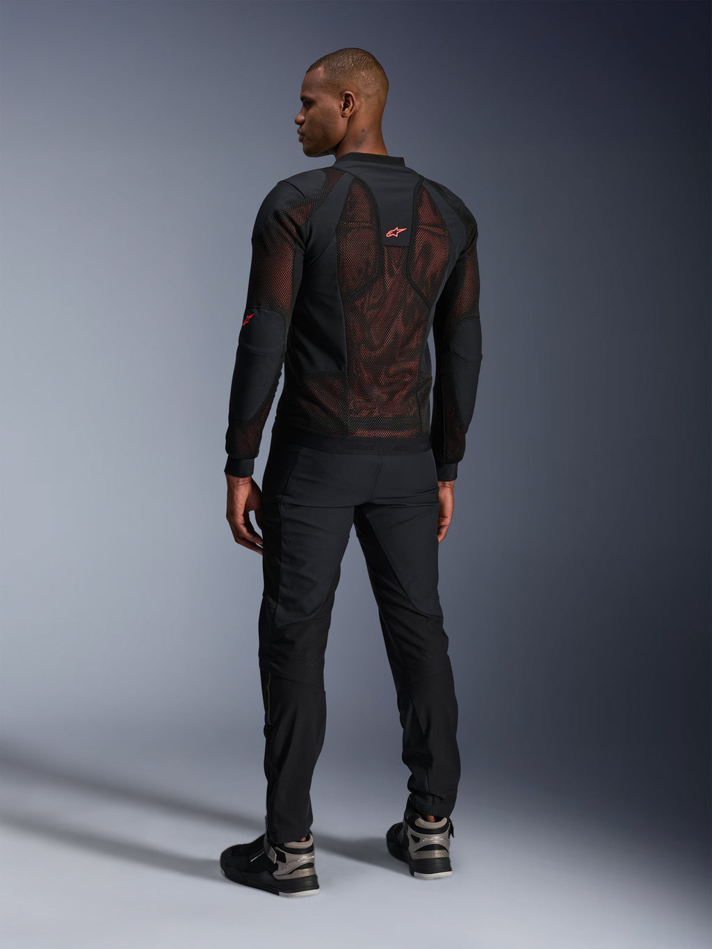 Alpinestars Troop-Air Pro-jacka i Black Red Fluo, ryggvy på en modell, med en stor ryggpanel i 2D-tech mesh med synlig röd insida, kombinerad med svarta tekniska körbyxor och svartgrå stövlar, modern urban design för en bepansrad motorcykeljacka.