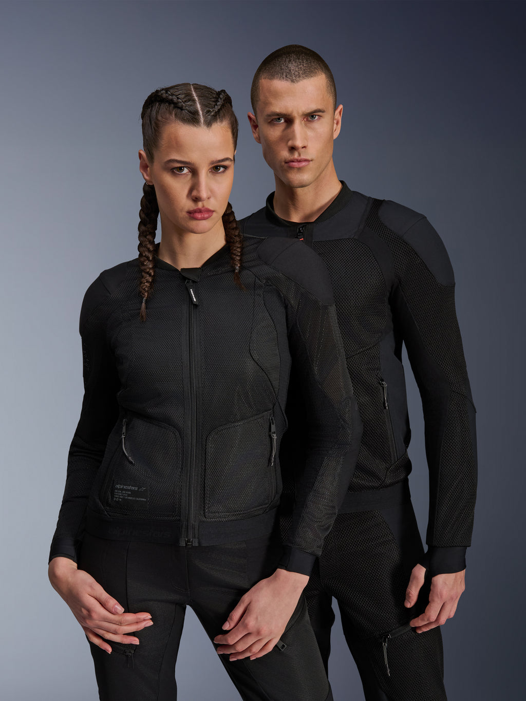 Man and woman wearing Alpinestars Troop-Air and Stella Troop-Air Jackets, black technical mesh motorcycle jackets, featuring 2D tech mesh structure for ventilation and reinforced panels, contemporary urban riding apparel.
