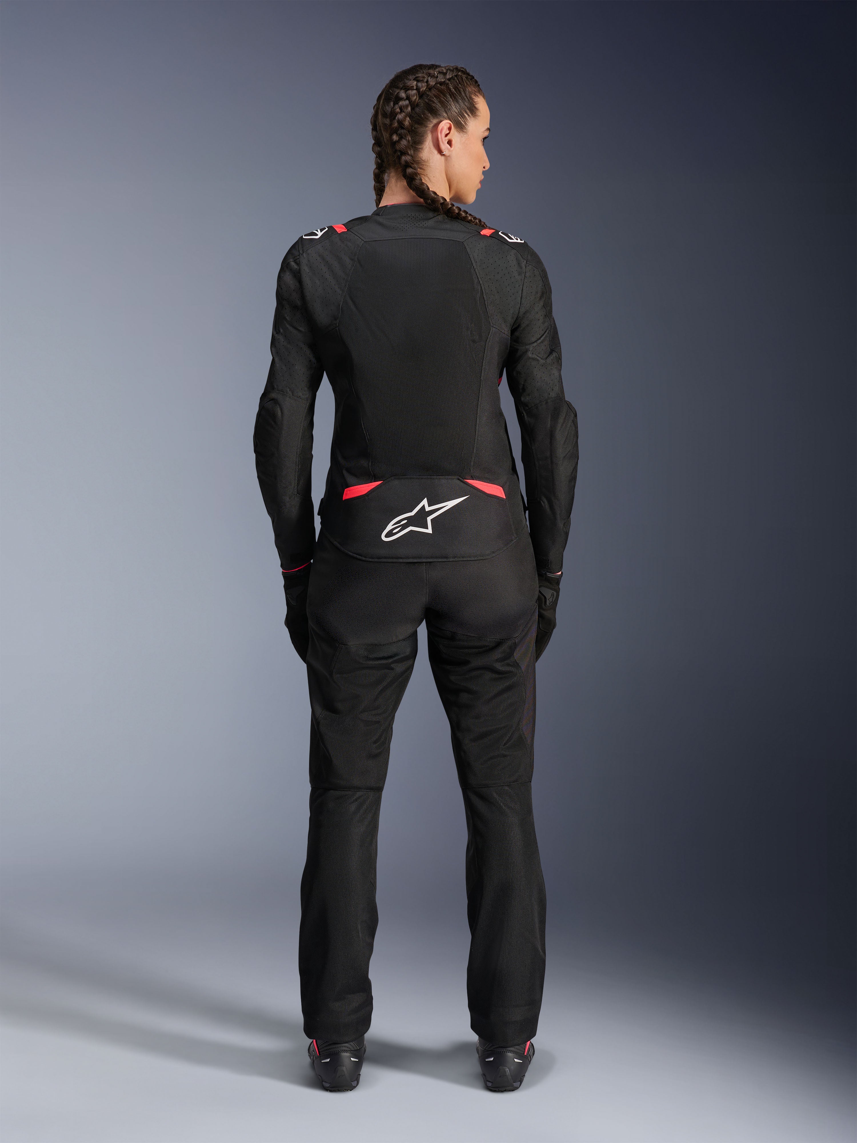 Alpinestars Stella T-SPS Air V2-jacka, motorcykeljacka i textil för dam, svart och Diva Pink, vy bakifrån på modell med flätat hår, med stora meshpaneler för ventilation, vit Astars-logotyp på nedre ryggen, matchad med svarta mc-byxor