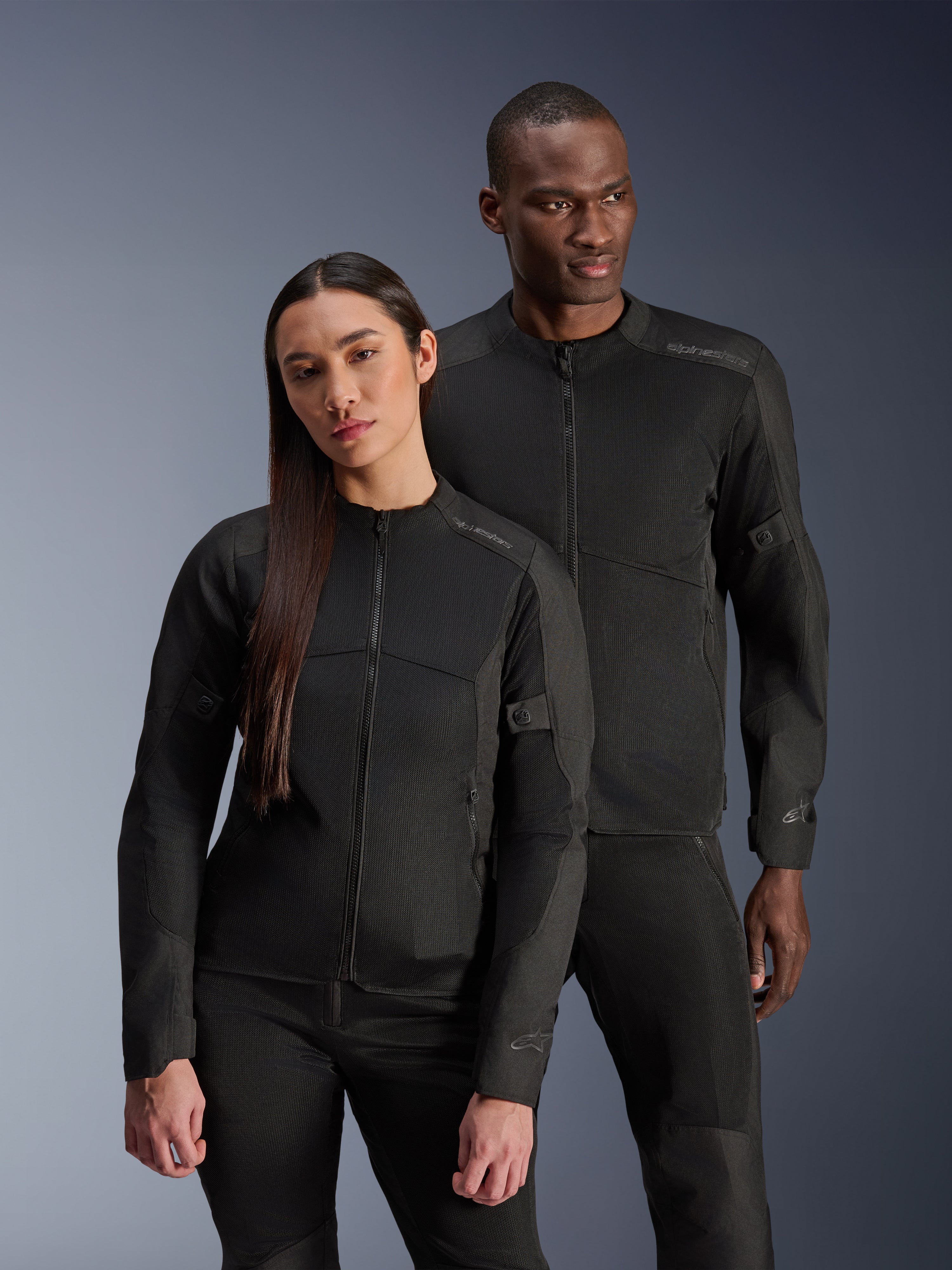 A man and a woman wearing the Alpinestars C-1 Air Jacket and Stella C-1 Air Jacket, matching black urban textile motorcycle jackets with mesh panels for ventilation, paired with black riding pants for a clean, contemporary look.