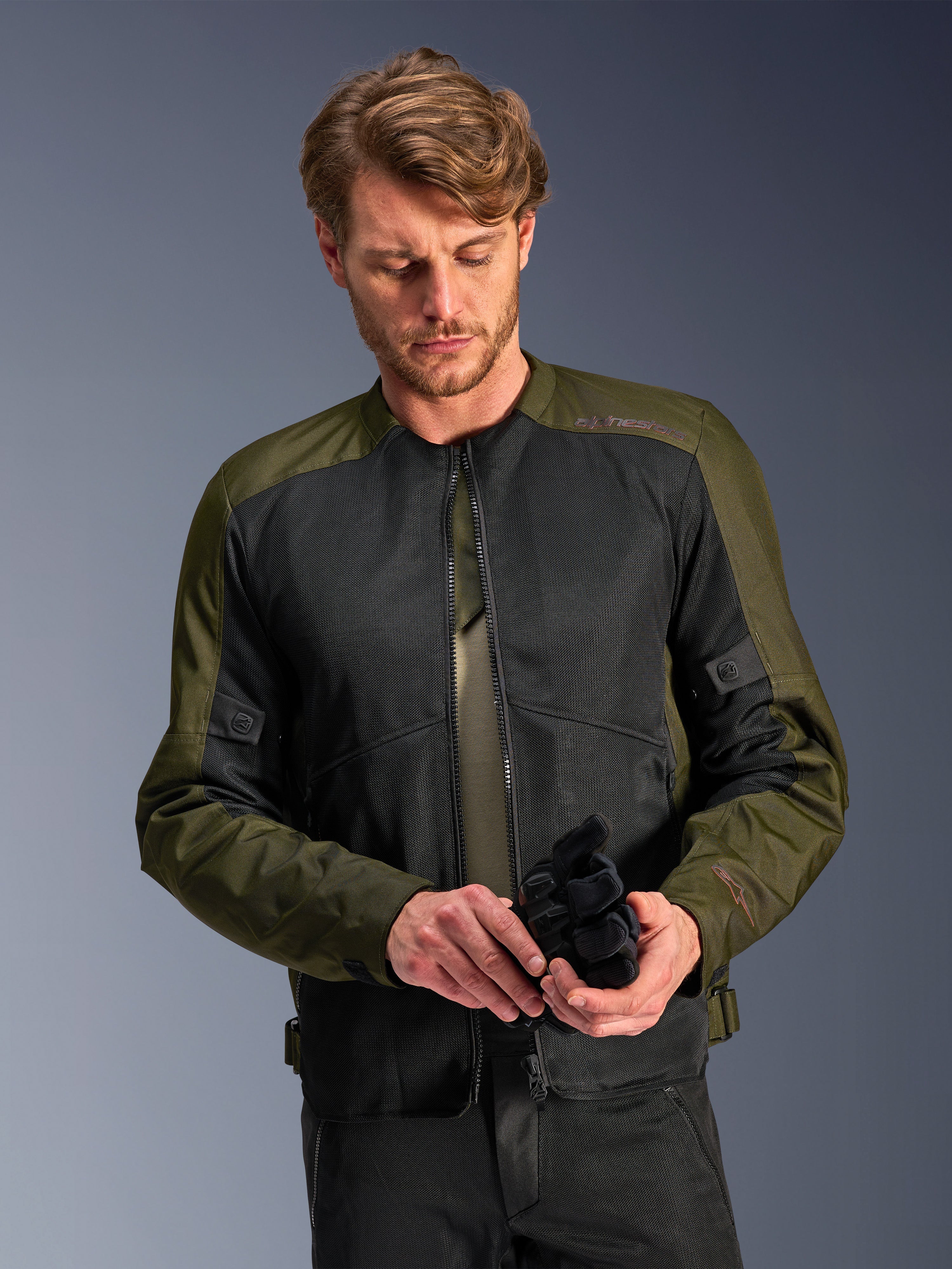 Man wearing the Alpinestars C-1 Air Jacket in Khaki, urban motorcycle jacket with olive green textile and black mesh panels, looking down while holding black motorcycle gloves, clean and contemporary style for road riding.