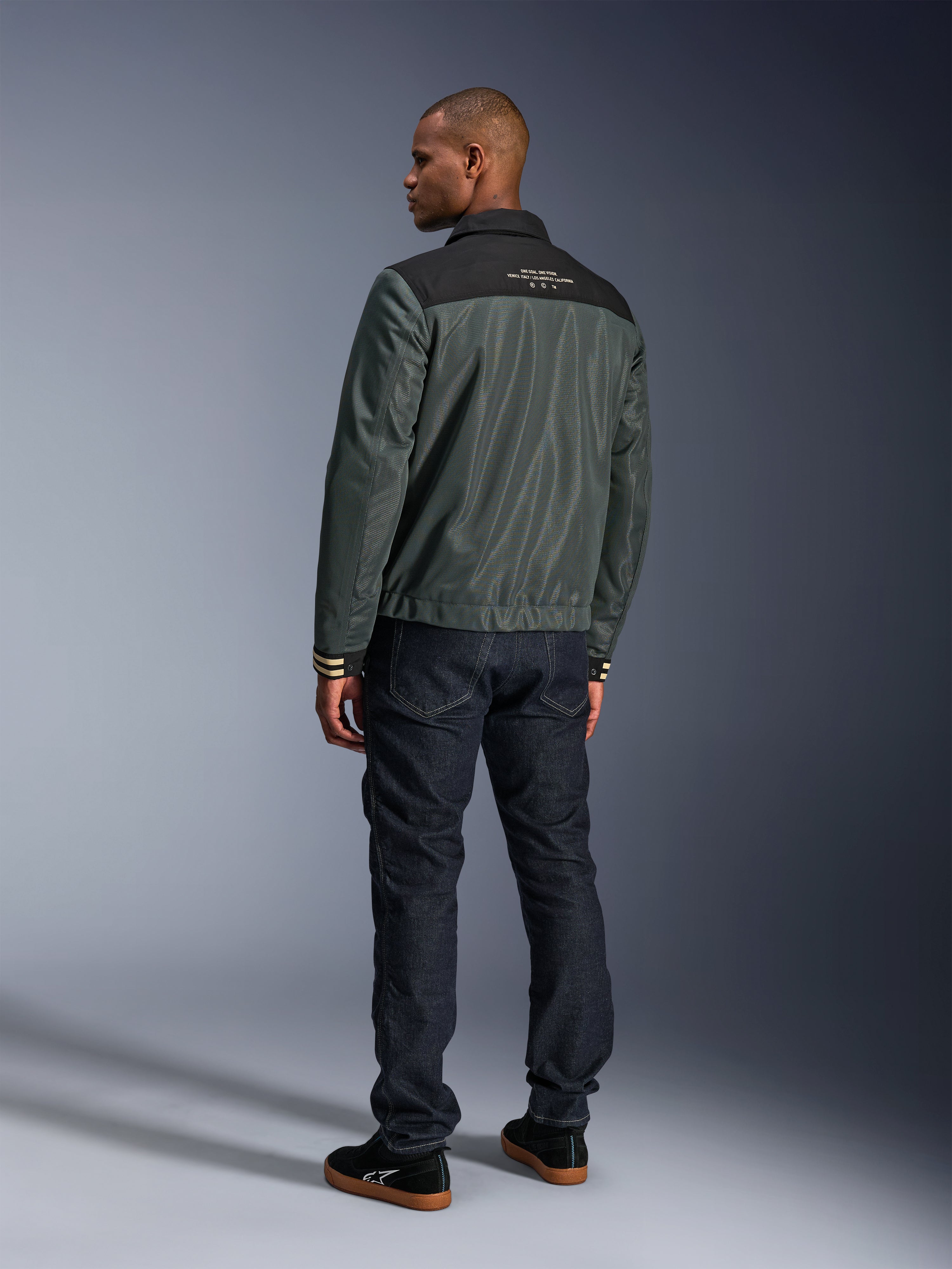 Alpinestars Ignite Air Jacket in Urban Gray Black Blue, rear view on model, featuring a mesh body with a black shoulder yoke and white text, striped cuffs, paired with dark denim jeans and black sneakers, urban lifestyle motorcycle apparel