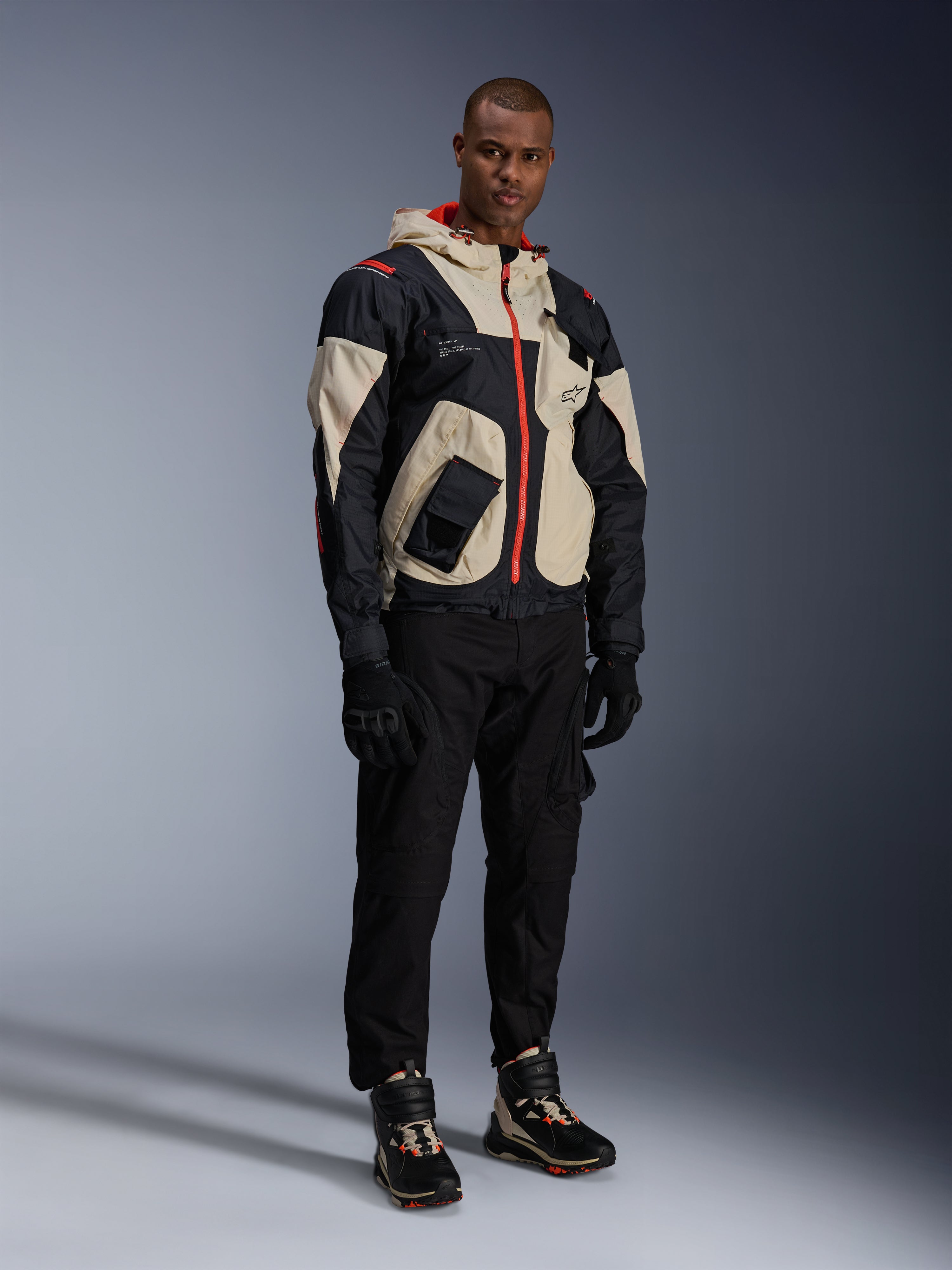 Man som står i komplett urban motorcykelutrustning, med Alpinestars Speedstreet Shoes i Black Almond Hot Orange och en Mogress Air Jacket i svart och krämfärgat, matchande svarta byxor och handskar, streetwear-inspirerad skyddsutrustning för stadskörning